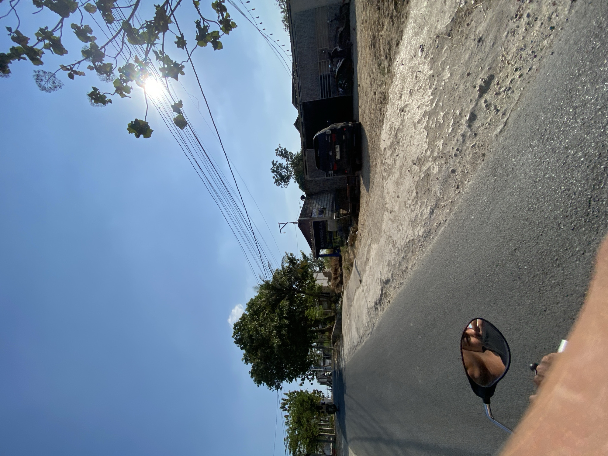 The image shows a perspective from someone riding a motorcycle on an asphalt road in an outdoor, rural setting under a clear blue sky. The sun is shining brightly, creating some lens flare and highlighting the leaves of a tree branch in the upper right corner. On the right side of the road, there are several buildings with corrugated roofs, a black SUV, and a motorcycle parked. The buildings appear to be low-rise structures with some signs of wear and tear. On the left side of the road, there are more trees and some structures, with another motorcycle visible in the distance. The foreground shows the rider's hand and arm and a reflection of their face in the motorcycle's rearview mirror. The overall impression is a sunny day in a village or small town.