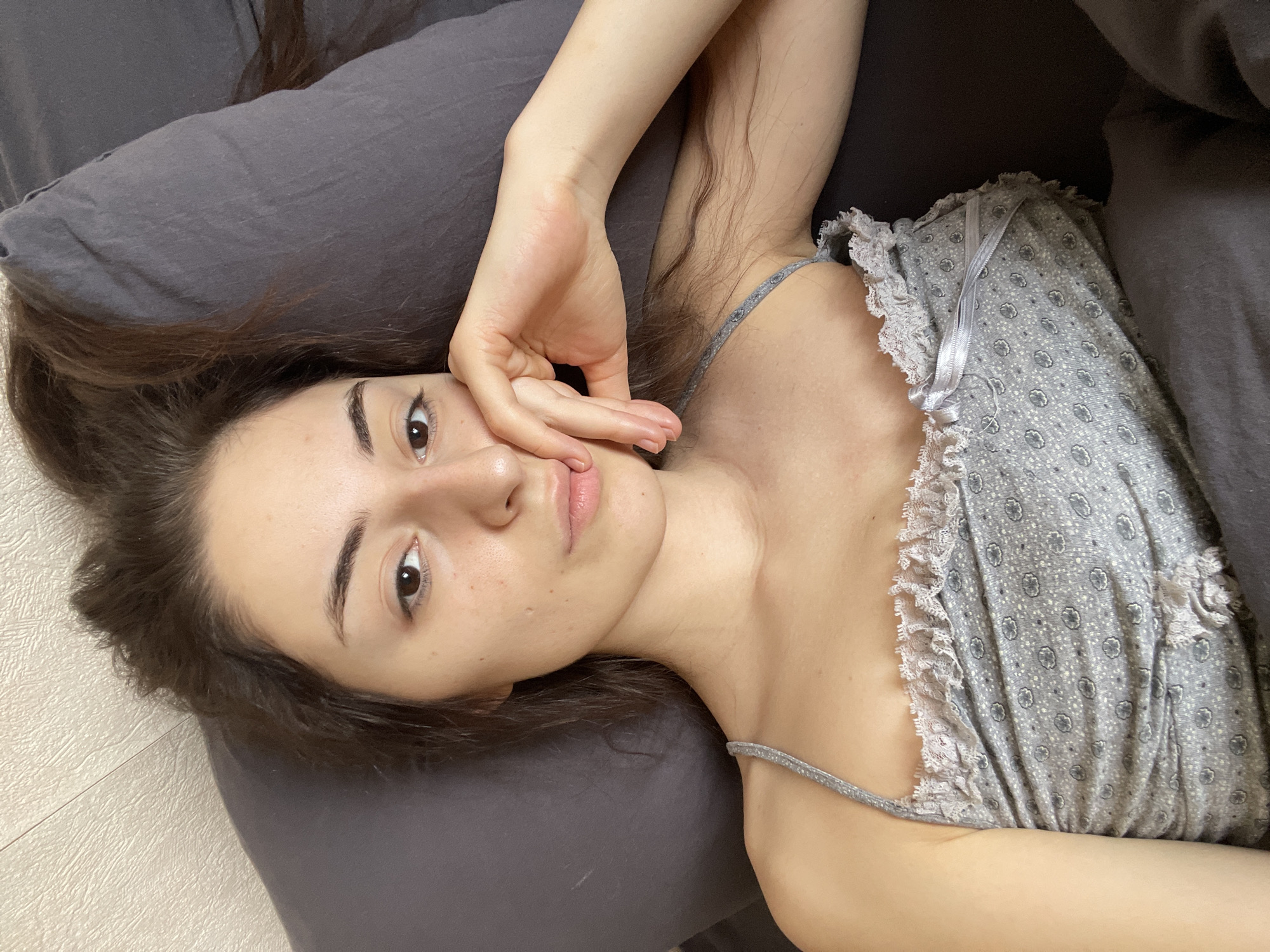 A young woman with brown hair is lying on a grey pillow in a bed covered with a grey blanket. She is wearing a grey and white patterned camisole with lace trim. Her right hand is touching her face. The location is not identifiable as Moscow, Russia.