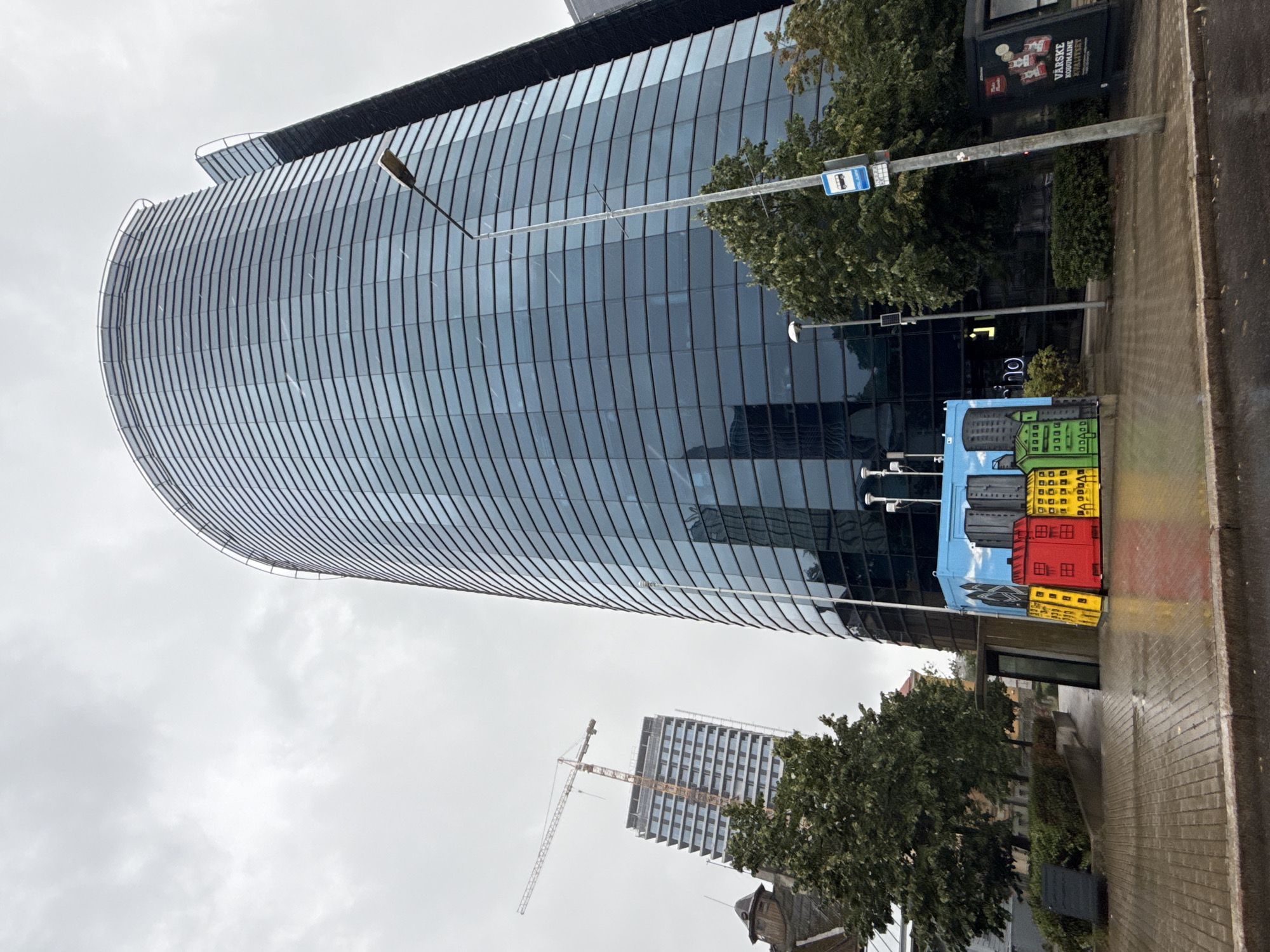 A contemporary cityscape in Tallinn, Estonia, features a tall, curved glass-fronted skyscraper, the Maersk Tower, dominating the scene. To the left, a partially constructed building with a crane signifies ongoing development. The sky is overcast, and the pavement appears wet, suggesting recent rain. In the foreground, a brightly painted, rectangular installation adorned with an urban mural of colorful buildings adds a vibrant touch. Streetlights and bus stop signage are visible, indicating a public urban area. Lush green trees and shrubs are interspersed throughout the urban landscape, providing a natural contrast to the modern architecture.