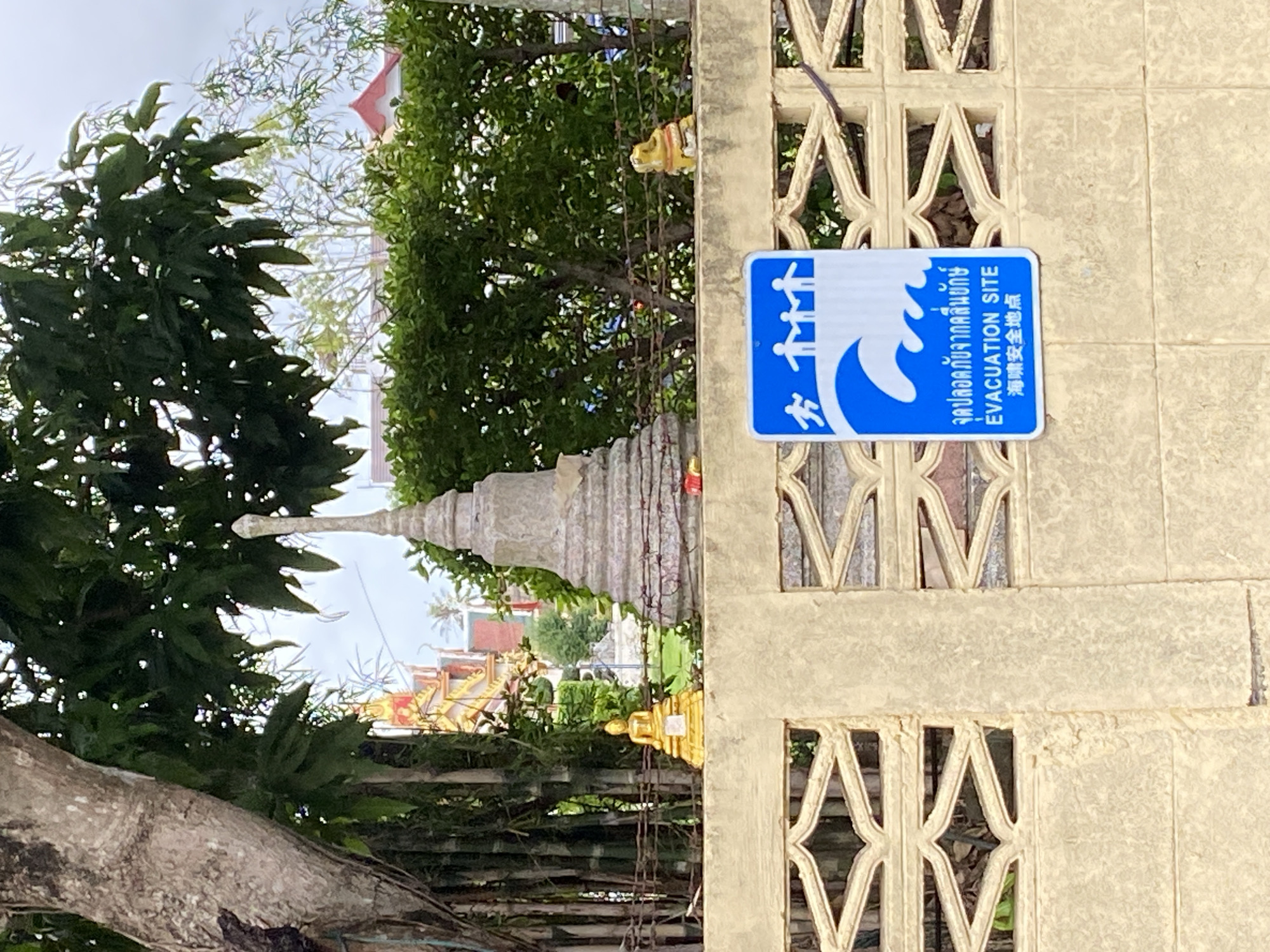 A sign that reads "Evacuation Site" in English, Thai, and Chinese is affixed to a concrete fence. The fence has a repeating geometric pattern. Behind the fence, there is a Buddhist temple. A tree partially obscures the view of the temple. The photograph was taken in Choeng Thale, Thailand.