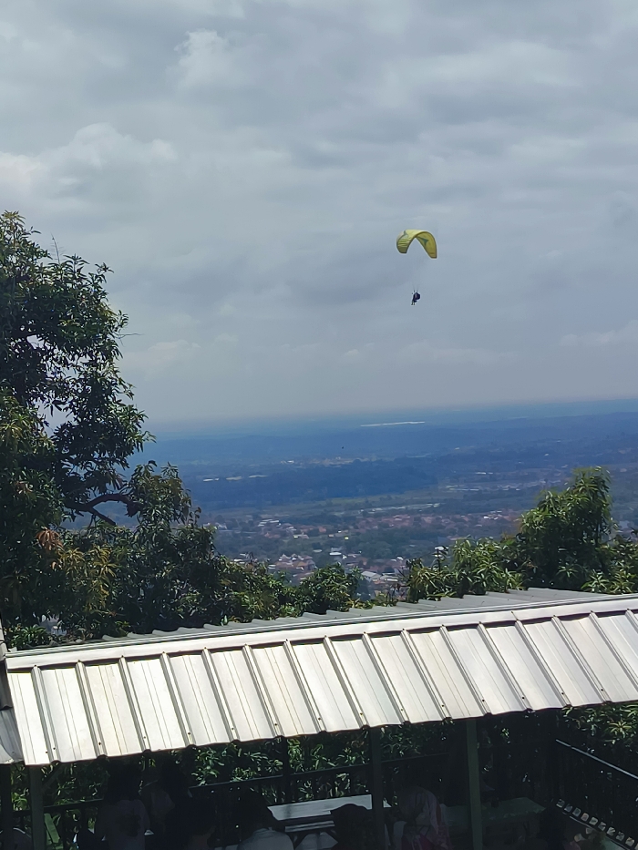A paraglider flies above a cityscape in Majalengka, Indonesia, located at latitude: -6.846, longitude: 108.204. The paraglider is a yellow and white canopy with a single figure visible inside. The sky is cloudy, with a blue haze visible in the distance. The scene is taken from a vantage point on a hill overlooking the city. A corrugated metal roofed structure sits in the foreground with people sitting underneath. The structure is made of wood and metal with a black railing. Green trees are on either side of the structure. In the distance, the city appears as a patchwork of buildings and greenery. The ground is visible in the distance, with a lot of green vegetation. The horizon is visible in the background, with a blue haze in the distance.