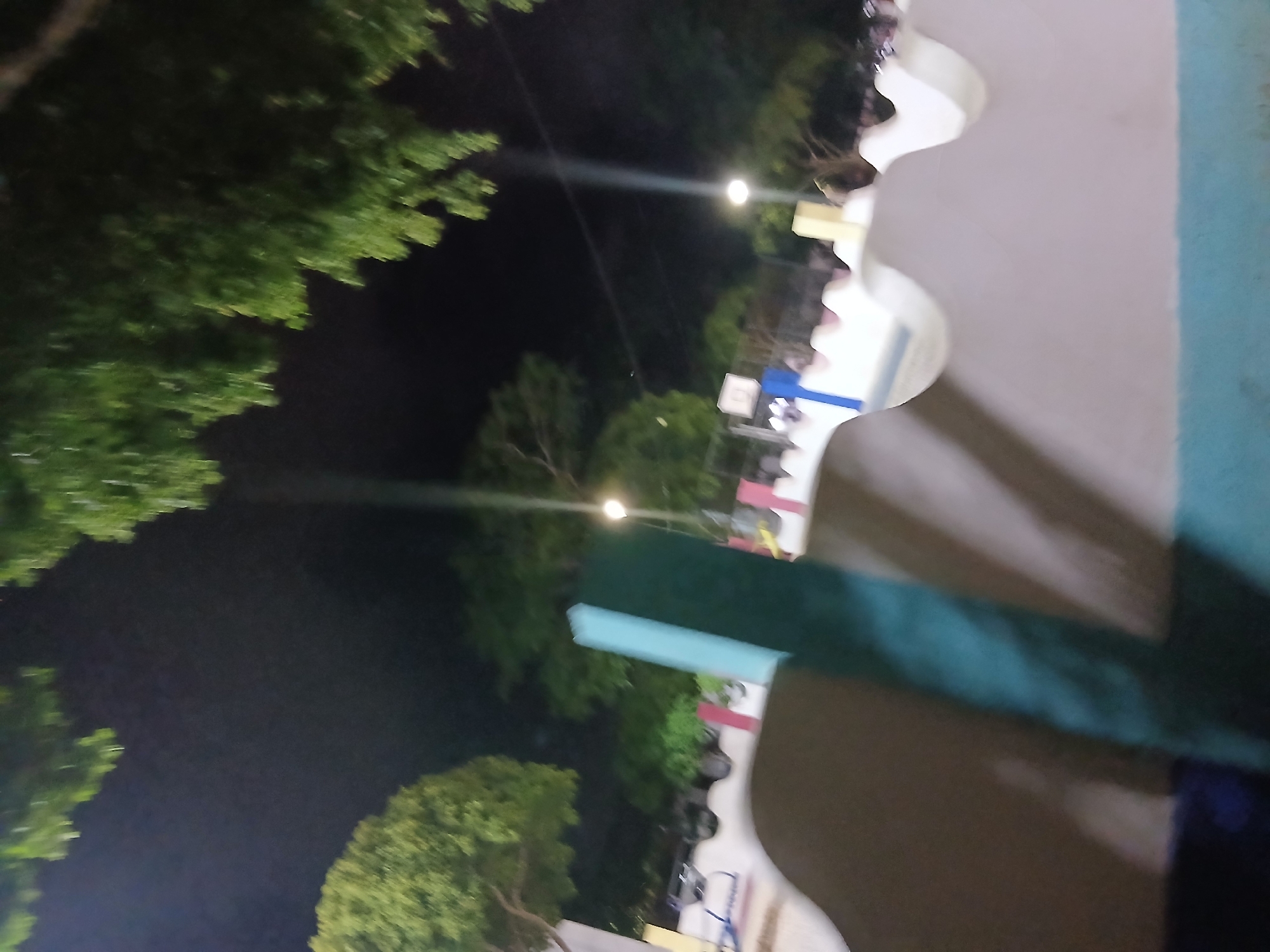 The image depicts a nighttime scene in a park in Santa Ana, Venezuela. The foreground is dominated by a dark, shadowed embankment with a light blue and white decorative edge. A prominent teal-colored vertical post stands in the middle ground. Beyond this, a playground area is visible with colorful structures, including a slide and a basketball hoop. Several parked cars are also visible in the background, illuminated by streetlights. Lush green trees with dense foliage frame the upper portion of the image, silhouetted against the dark night sky. The overall ambiance is that of an outdoor public space at night, with artificial lighting casting shadows and highlighting the playground equipment.