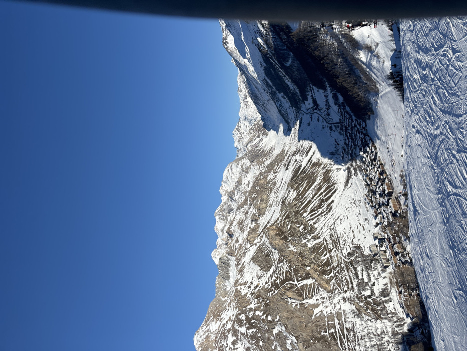 This vibrant, high-angle outdoor shot captures a majestic snow-covered mountain landscape under a brilliant, clear blue sky. The image appears to be taken from a ski lift, gondola, or elevated viewpoint, with a dark horizontal bar visible at the top (which would be a vertical bar on the left if rotated correctly to landscape orientation), suggesting part of the viewing structure.
The scene showcases the rugged beauty of the French Alps, specifically Val-d'Isère. Dominating the frame are towering, snow-dusted mountains, with exposed rocky faces revealing intricate geological patterns. Patches of dark evergreen trees cling to the steeper slopes, contrasting with the white snow and grey rock.
Towards the right side of the image, a wide, meticulously groomed ski piste is visible, crisscrossed with numerous ski tracks, indicating active skiing. Nestled further down, what appears to be a village or cluster of buildings, likely traditional chalets or resort facilities, is visible, their roofs blanketed in snow. Other ski infrastructure, such as potential lift towers, can be faintly discerned in the distance.
The bright sunlight and clear blue sky suggest a perfect winter day, ideal for outdoor activities like skiing. The overall atmosphere is one of serene grandeur and the excitement of a popular ski destination. No discernible text is visible in the image. People are implied by the ski tracks and resort setting but are not individually visible.