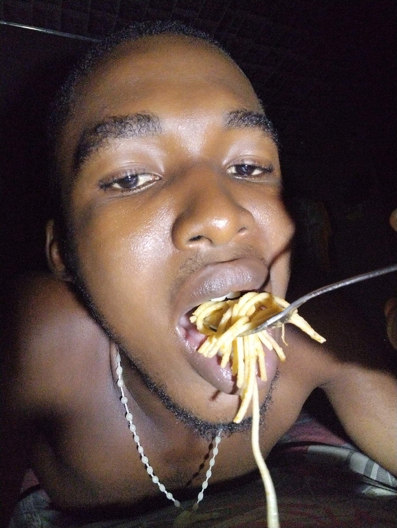 A close-up, low-light photograph shows a dark-skinned male in Ihiagwa, Nigeria, in the process of eating. He is depicted from the chest up, with his face prominently featured. A fork, held in his right hand, is positioned to enter his mouth, laden with strands of spaghetti or noodles. His eyes are open and directed towards the camera, while his mouth is slightly open around the food. He has short, dark hair and light facial hair on his chin and jawline. His skin appears moist or oily, with highlights reflecting artificial light on his forehead, nose, and cheeks. Around his neck, a necklace made of small, light-colored beads is visible. The background is dark and indistinct, suggesting an indoor setting, likely at night or in a dimly lit room. No text is visible in the image.