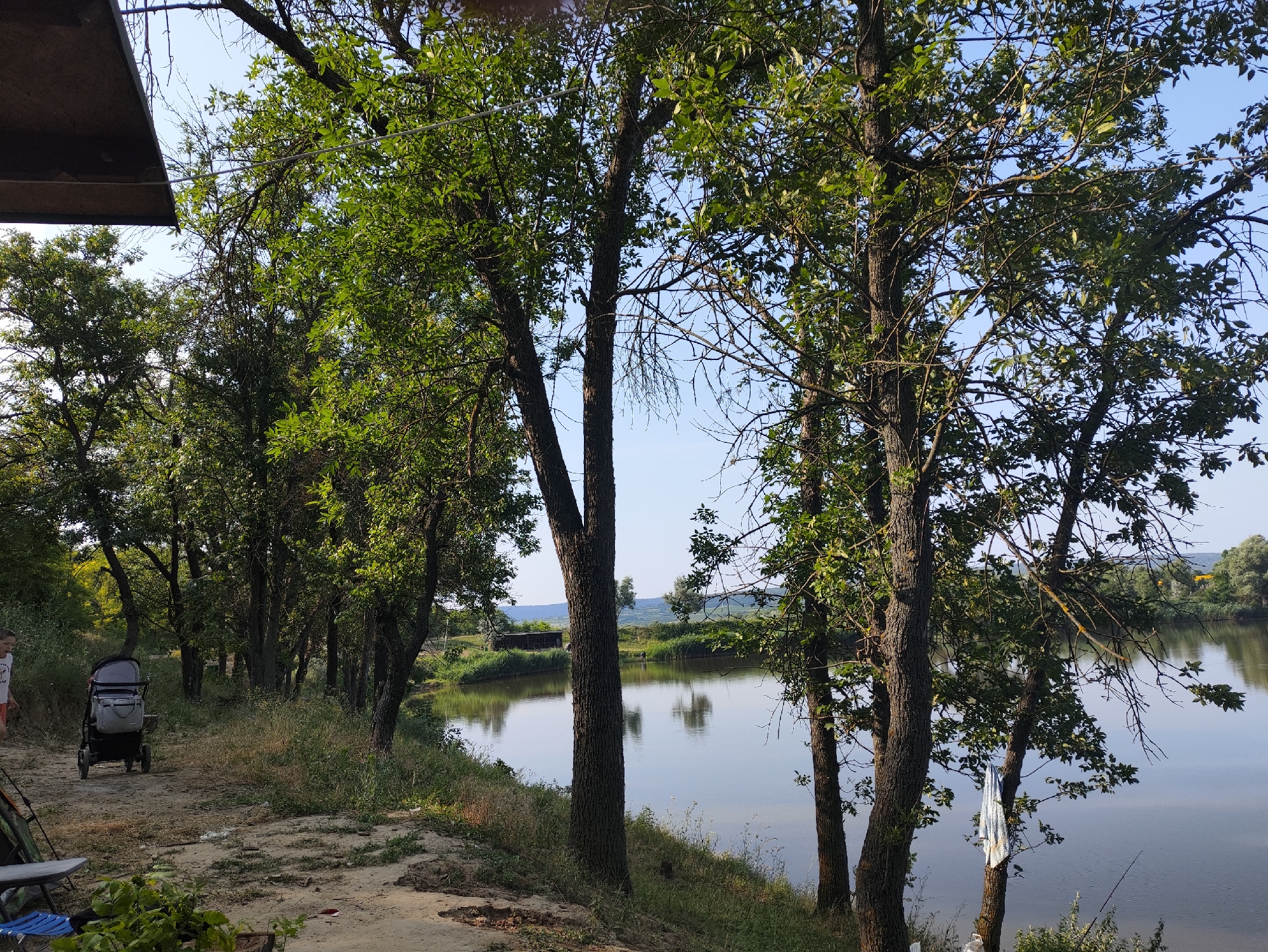 The image depicts a serene natural scene, likely in Găleștii Noi, Moldova, featuring a tranquil river or lake on the right side of the frame. Dense foliage from numerous trees lines the bank, with their reflections shimmering on the water's surface. A clear blue sky overhead suggests it is daytime. On the left bank, a sandy path winds along the water's edge. A child, wearing a white shirt and orange shorts, is seen walking away from the camera, pushing a dark-colored stroller. In the foreground, to the left, a portion of a table and a blue folding chair are visible, indicating a potential picnic or resting area. The overall ambiance is peaceful and rural.