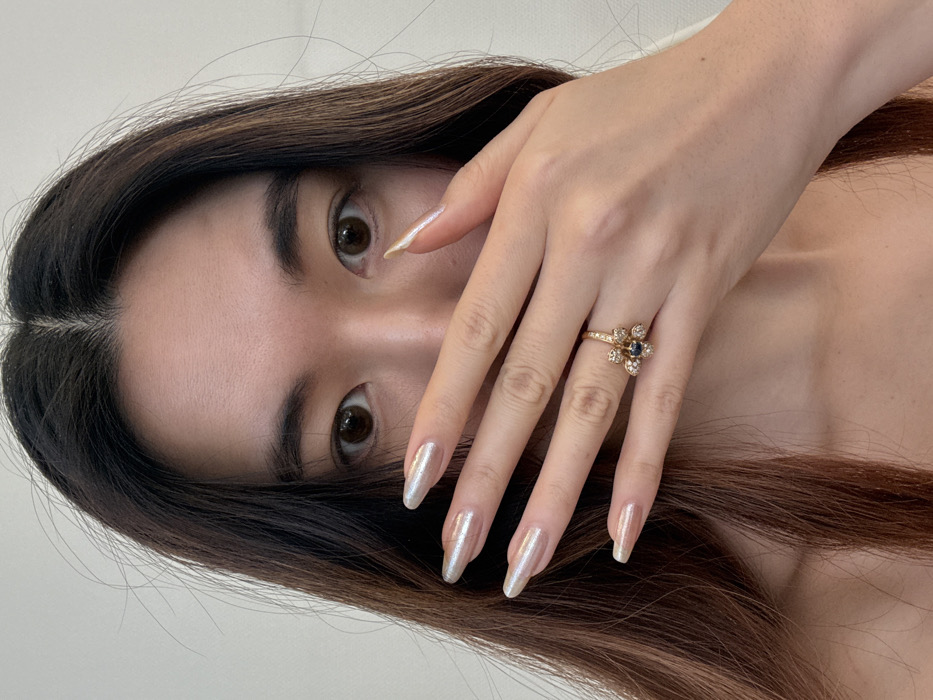 A close-up, head-and-shoulders portrait of a person, likely female, with long, dark brown hair parted centrally. The individual's dark brown eyes are open and directed towards the viewer. The left hand is raised, partially covering the lower half of the face, specifically the nose and mouth area. The fingers are extended, displaying long, almond-shaped nails with an iridescent, pearlescent light finish. A gold-toned ring, featuring a central dark blue or black gem surrounded by smaller clear stones arranged in a floral pattern, is worn on the fourth finger of the left hand. Exposed skin on the upper chest and shoulders indicates a garment with an open or wide neckline. The background is a uniform, plain light color, suggesting an indoor setting. This scene is located in Arcueil, France.