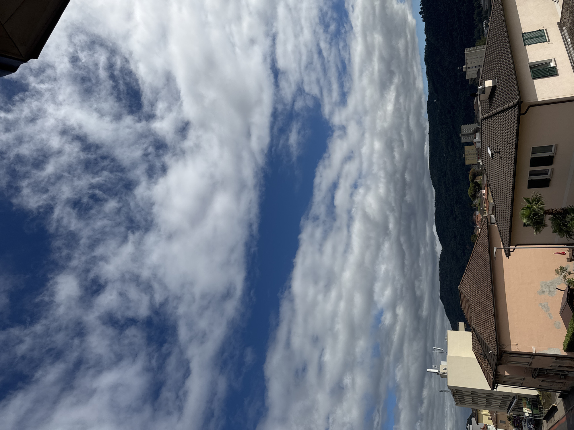 The image shows a view of Balerna, Switzerland, from a slightly elevated vantage point. The sky is a dominant feature, filled with a mix of bright blue patches and white, wispy clouds, with some darker, heavier clouds present towards the horizon. Below the sky, a dark, tree-covered hill forms the backdrop of the scene. In the mid-ground, several buildings are visible. Closer to the foreground, there are tiled rooftops of residential buildings. One prominent building on the left is a light-colored, multi-story structure with antennas on its roof. To its right, a pale pink building with a tiled roof is visible. Further to the right, another residential building with a dark tiled roof and green shutters is partially visible. The environment appears to be urban or suburban, with evidence of housing and infrastructure. No people are depicted in the image.