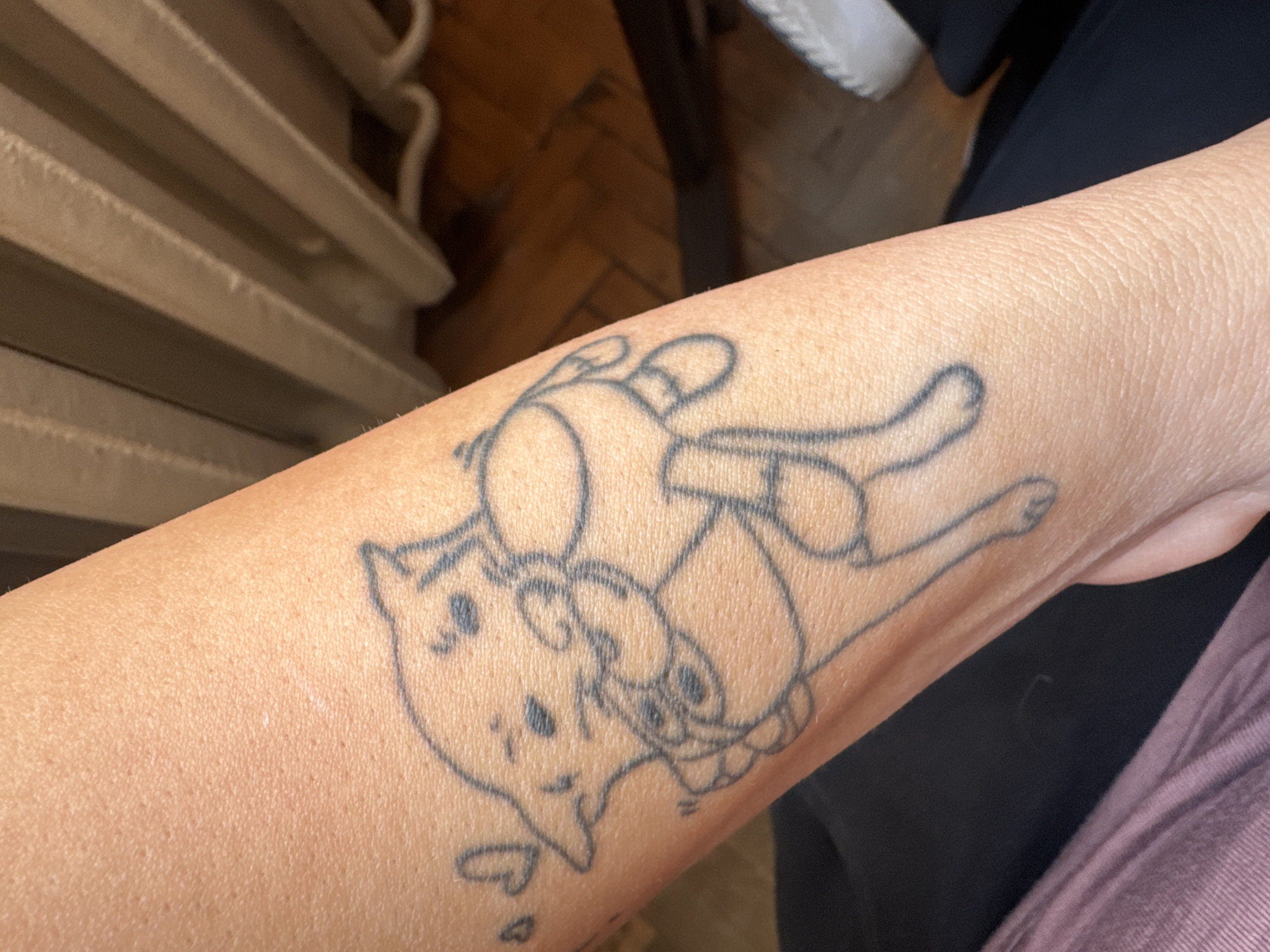 A close-up shot shows a person's forearm with a black line tattoo of two cartoon characters. The background is blurred, showing a radiator and some clothing. The location is unknown.