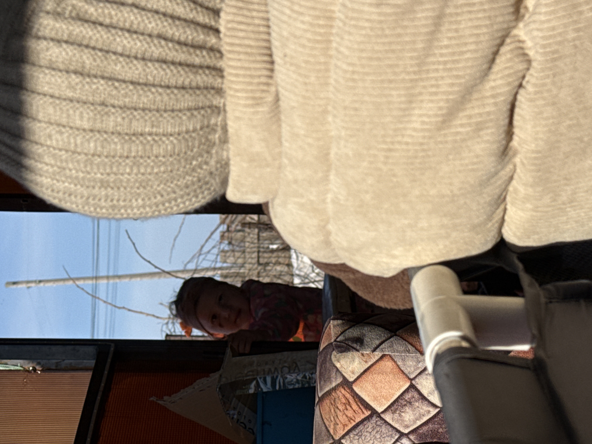 A low-angle shot from within a structure looks out to the sky and a young child. The child peers in, framed by a window or opening. Outside, bare tree branches are visible against a blue sky. A utility pole stands in the distance. Inside, a person wearing a light-colored, ribbed sweater or cardigan is positioned above the viewer, partially obscuring the view. A patterned cushion and a portion of a black and white structure with a plastic pipe are also in the foreground. The location is Кипчак, Russia.
