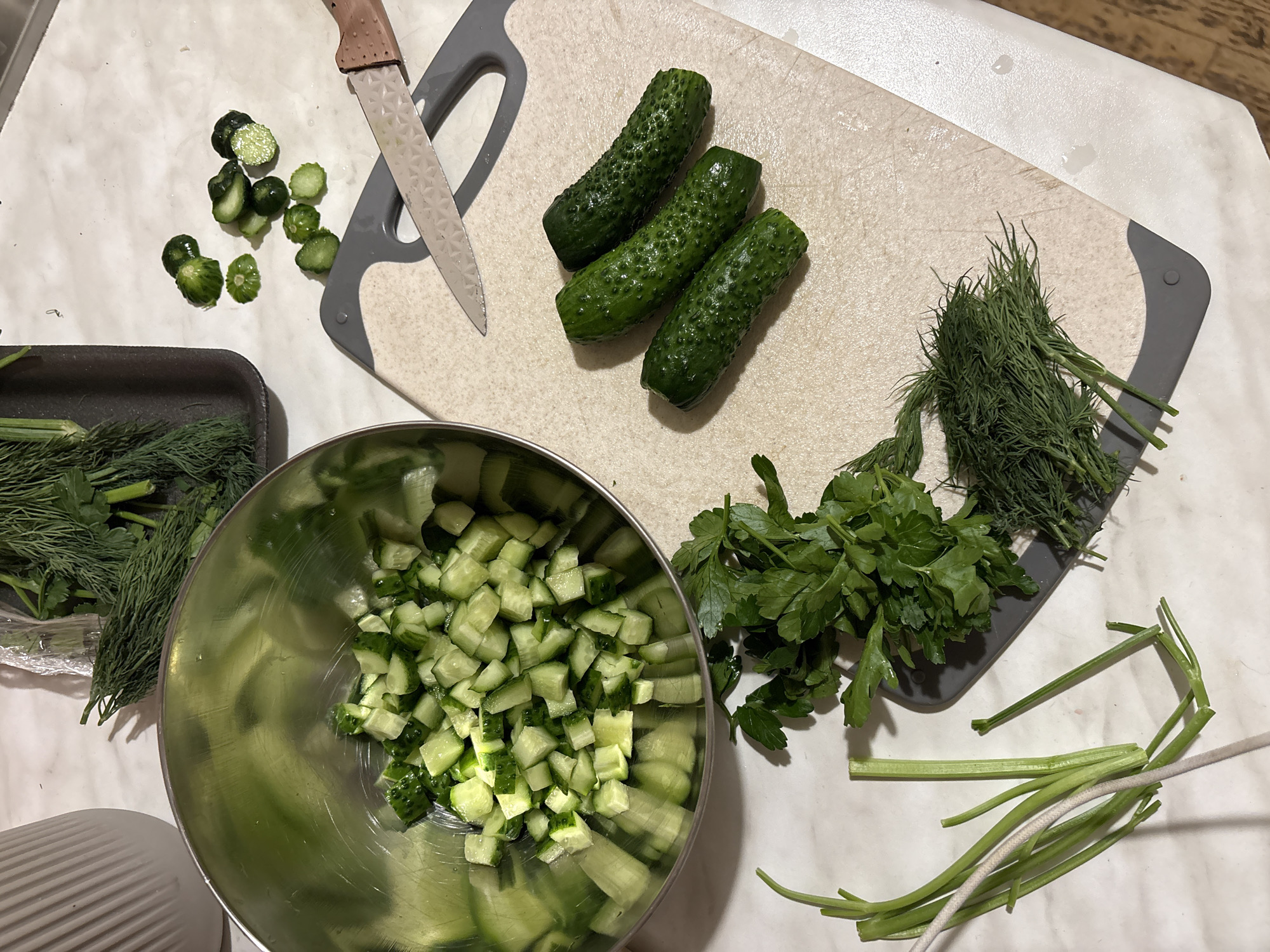The image shows a food preparation scene, likely in a domestic kitchen. There are three whole cucumbers on a cutting board, along with a pile of dill and parsley. A knife lies near some cucumber pieces. A bowl is mostly filled with diced cucumbers. To the left, a container holds more dill and parsley. Some stray parsley stalks and a white cord are on the surface. The scene is not identifiable as specifically being in Moscow, Russia.