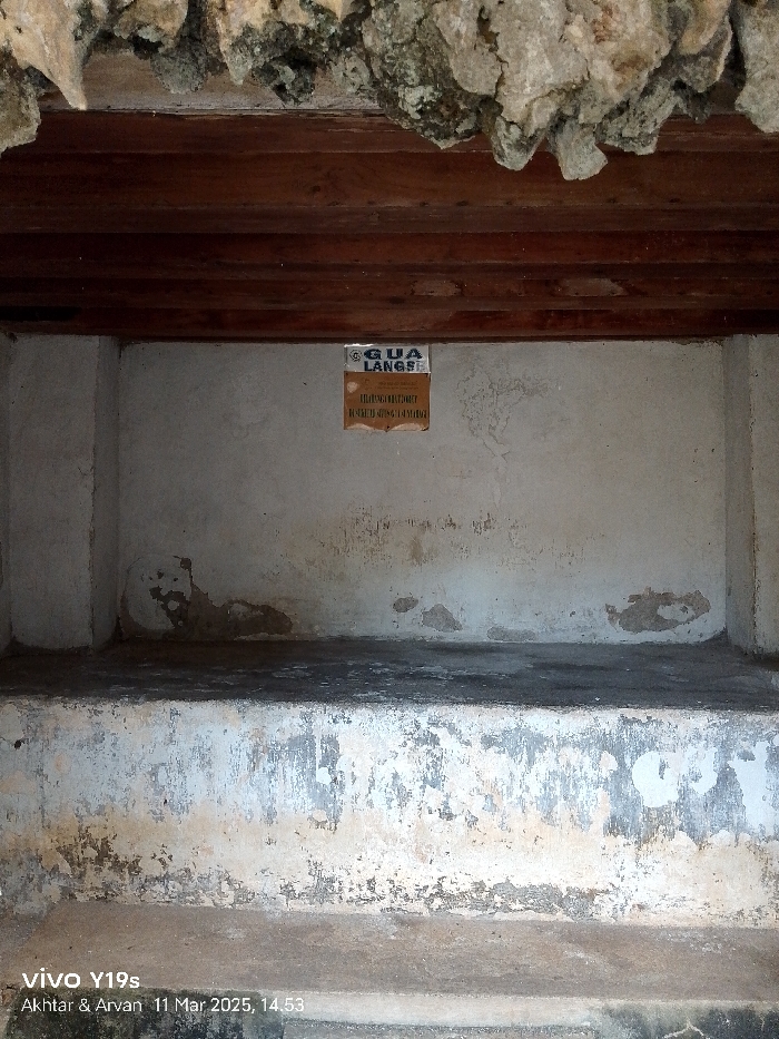 The image shows the interior of a small, rectangular, concrete room with a wooden ceiling. The room is empty except for a rectangular, yellow sign on the wall. The sign reads "GUA LANGS," "TIDAK DI PERBOLEHKAN," and "MEMBUANG SAMPAH DI TEMPAT INI." The walls and ceiling are weathered and have peeling paint. The coordinates (-6.736, 108.544) place the location of the room in Cirebon, Indonesia.