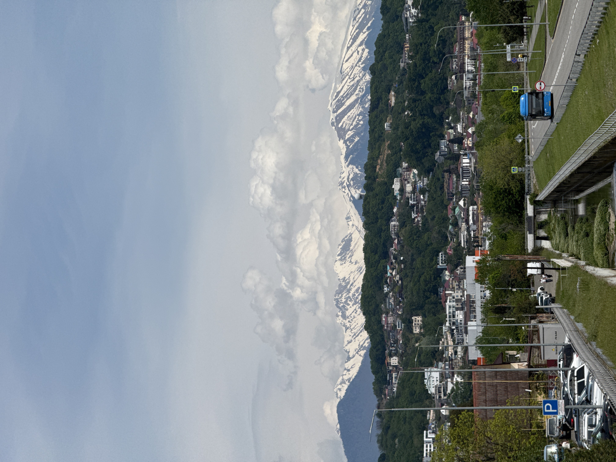 The image presents a view of Sirius, Russia. The scene includes a cityscape at the base of a mountain range with partial snow cover. Buildings of varying sizes and architectural styles populate the hillside. A road runs along the right side of the image, with a blue vehicle visible on it. A sidewalk and a metal railing border the road. A "P" parking sign is visible among parked vehicles on the foreground. The sky is partly cloudy.