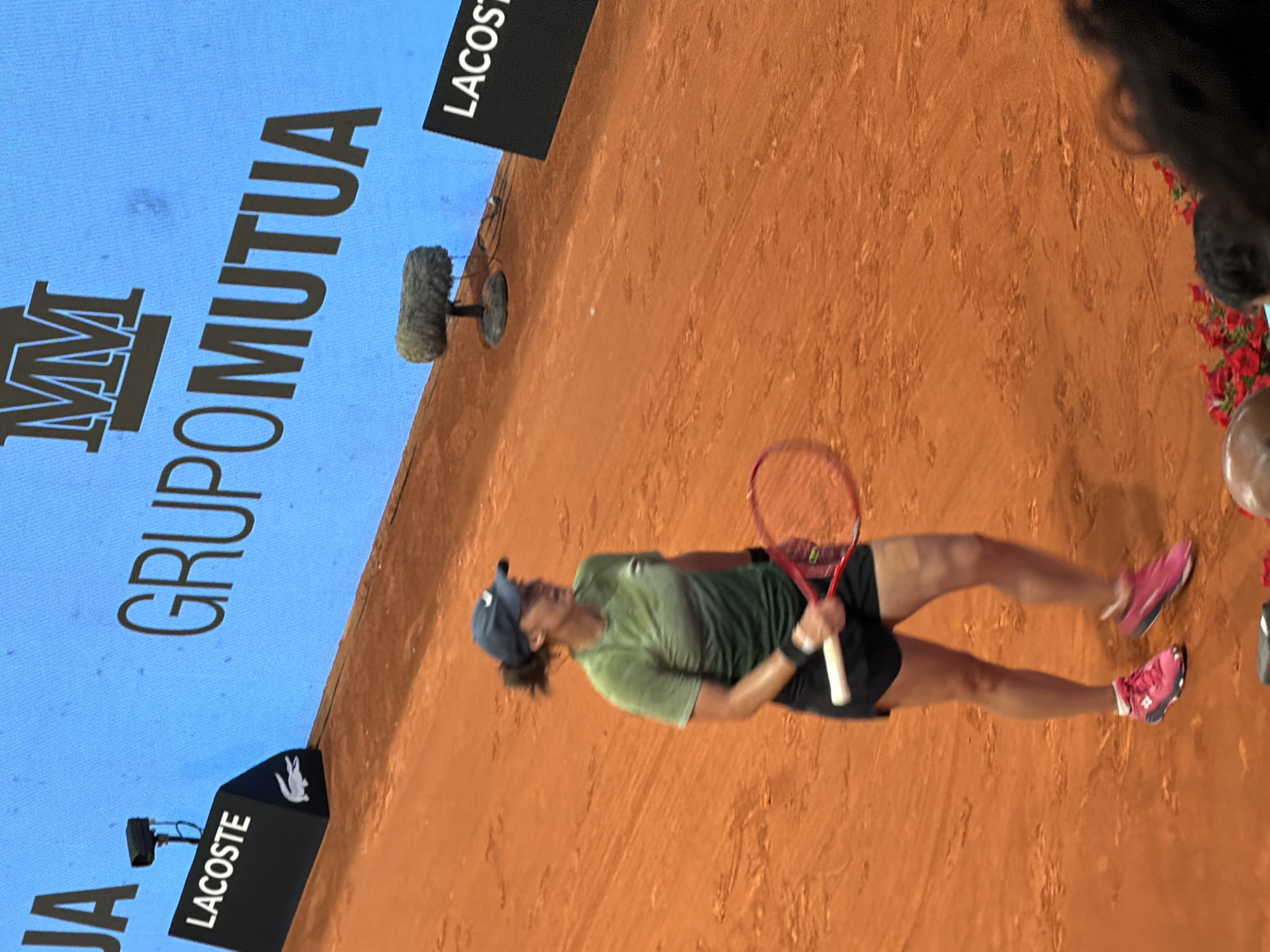 A tennis player wearing a green shirt, black shorts, and pink shoes is playing on a clay court. She is holding a red and white tennis racket. In the background, there is a blue screen with the words "GRUPO MUTUA MM" printed on it. There are also advertisements for Lacoste. The location is Madrid, Spain.