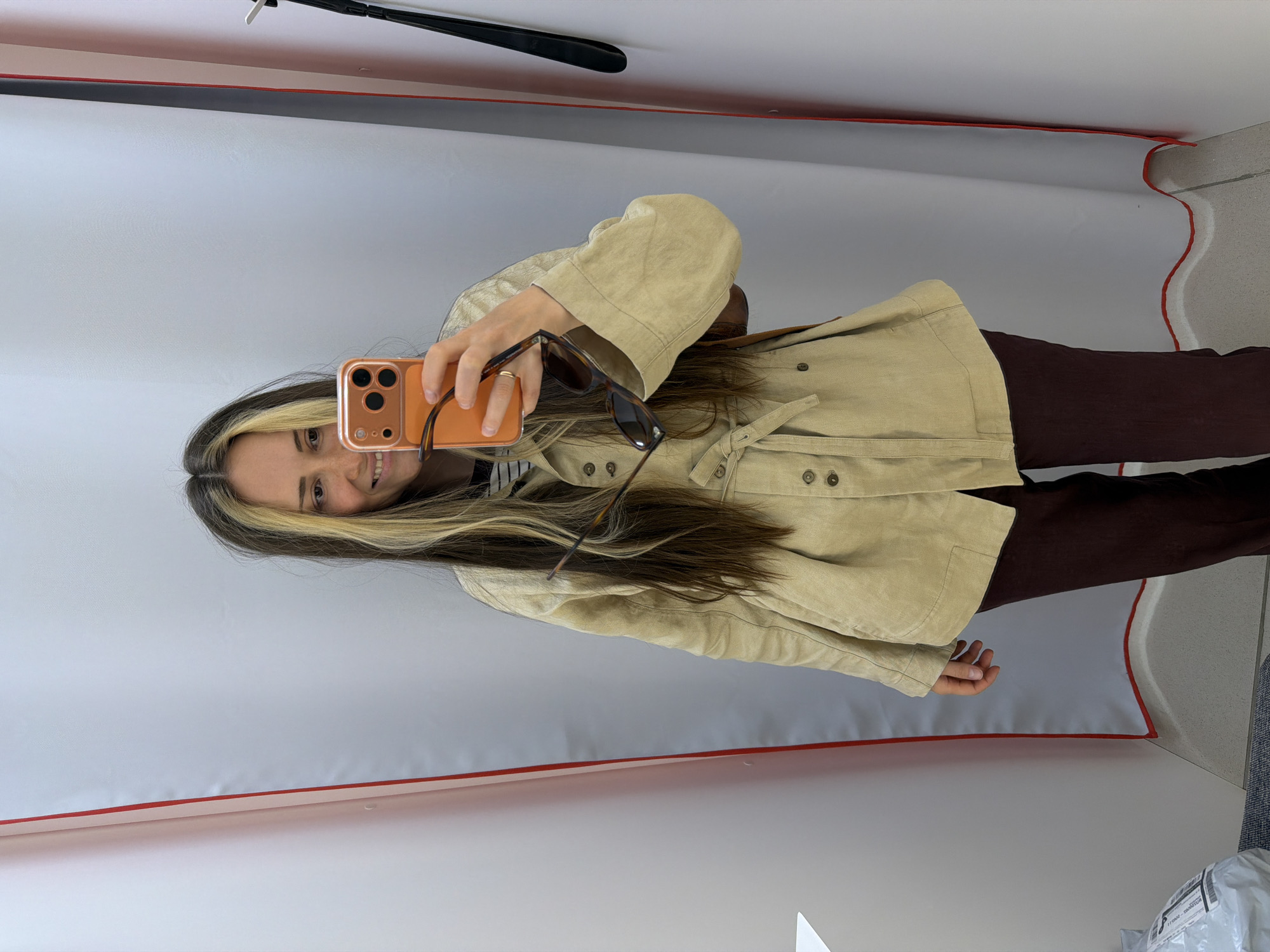 A woman is taking a selfie in what appears to be a fitting room. She is holding a phone with an orange case in her right hand, and a pair of sunglasses in her left hand. She is wearing a tan jacket with button details and brown pants. The fitting room is surrounded by white fabric with a red border. In the upper left corner of the frame, a black hanger is visible, attached to a bar. The floor is partially visible with what looks like a package. The location is unknown and there is no indication that the location is in Kamyshin, Russia.