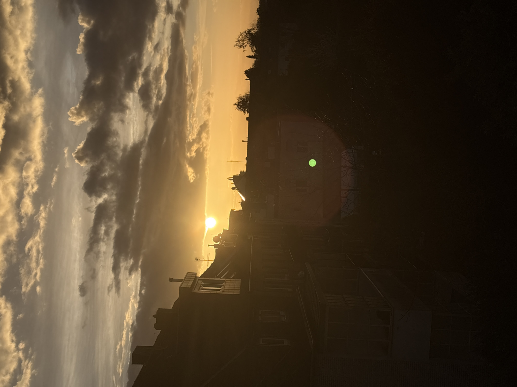 The image depicts a sunset scene in Mönchengladbach, Germany. The sun is low on the horizon, obscured by a layer of clouds that are illuminated with golden light. Silhouetted buildings are visible in the foreground, including residential structures with visible windows, chimneys, and rooftop antennas. A row of trees lines the upper part of the skyline, also appearing silhouetted. The sky is filled with varying shades of orange and gray, indicating a combination of clear sky and cloud cover. A lens flare is visible, caused by direct sunlight hitting the camera lens.