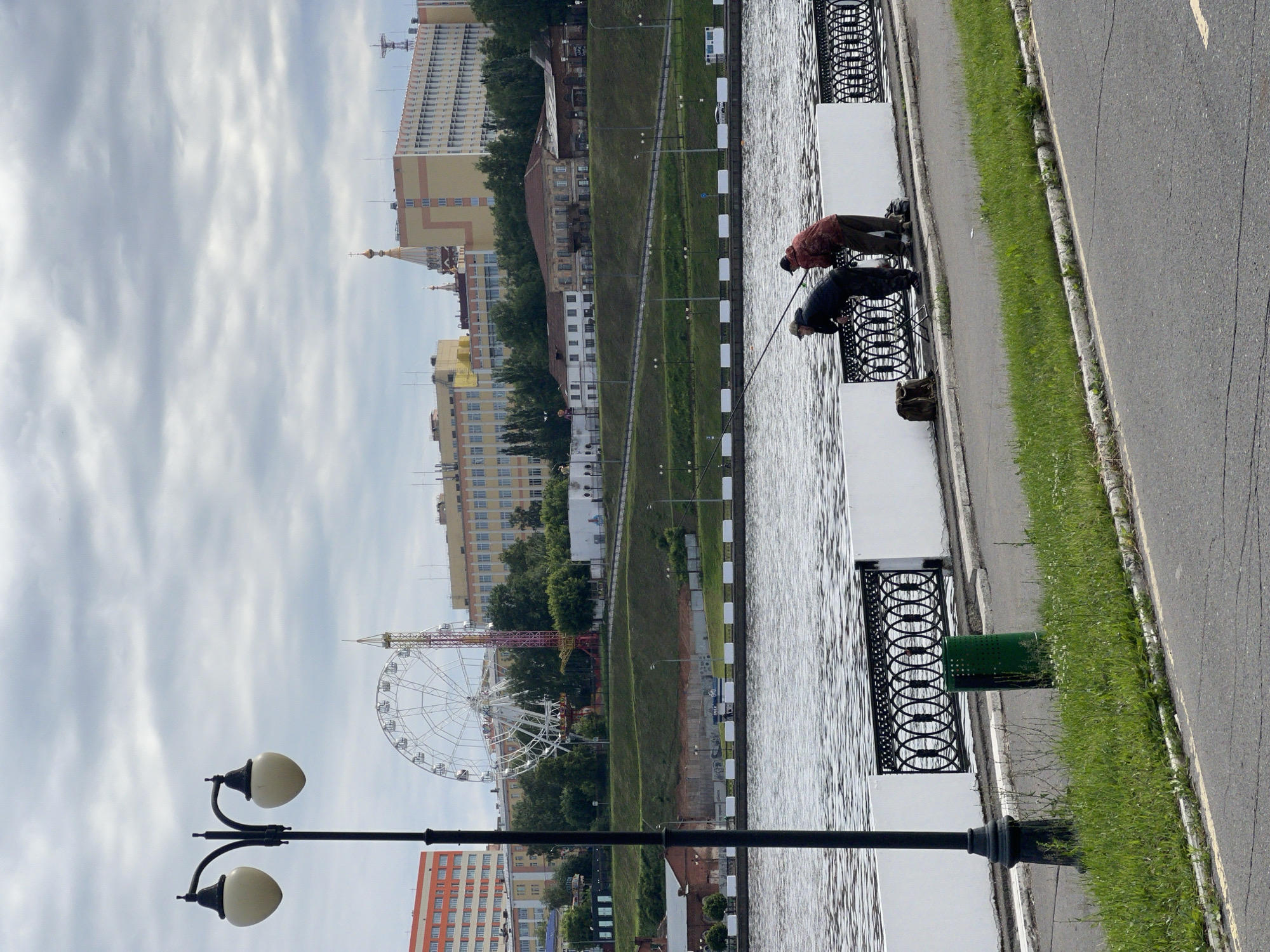 This image depicts a daytime urban scene along a waterfront in Izhevsk, Russia, under an overcast sky. In the foreground, a black, ornate street light with two white globe fixtures stands to the left, with a green cylindrical trash bin positioned below it. An asphalt path with a yellow centerline shows cracks. A strip of grass separates the path from a white concrete embankment wall topped with black, ornate metal railings.
Two adult males are positioned at the railing in the right-center, leaning over the wall and actively engaged in fishing. Both hold fishing rods extending into the choppy water. The male on the left wears a dark jacket and pants, while the male on the right wears a reddish-brown jacket and brown pants. Fishing equipment, including a small stool and bags, rests on the ground beside them.
Across the water, a green, grassy hillside rises, traversed by paved pathways and illuminated by several lampposts. Various buildings are situated on and behind the hillside. Notable structures in the background skyline include a large white Ferris wheel, an adjacent tall amusement park ride (possibly a drop tower), and a cluster of multi-story urban buildings of varying colors (orange, yellow, light blue/grey). A prominent golden dome topped with a cross, indicative of an Orthodox church or cathedral, is visible amongst the buildings. The large Ferris wheel is identifiable as the "Wheel of Izhevsk," located in the Central Park.
