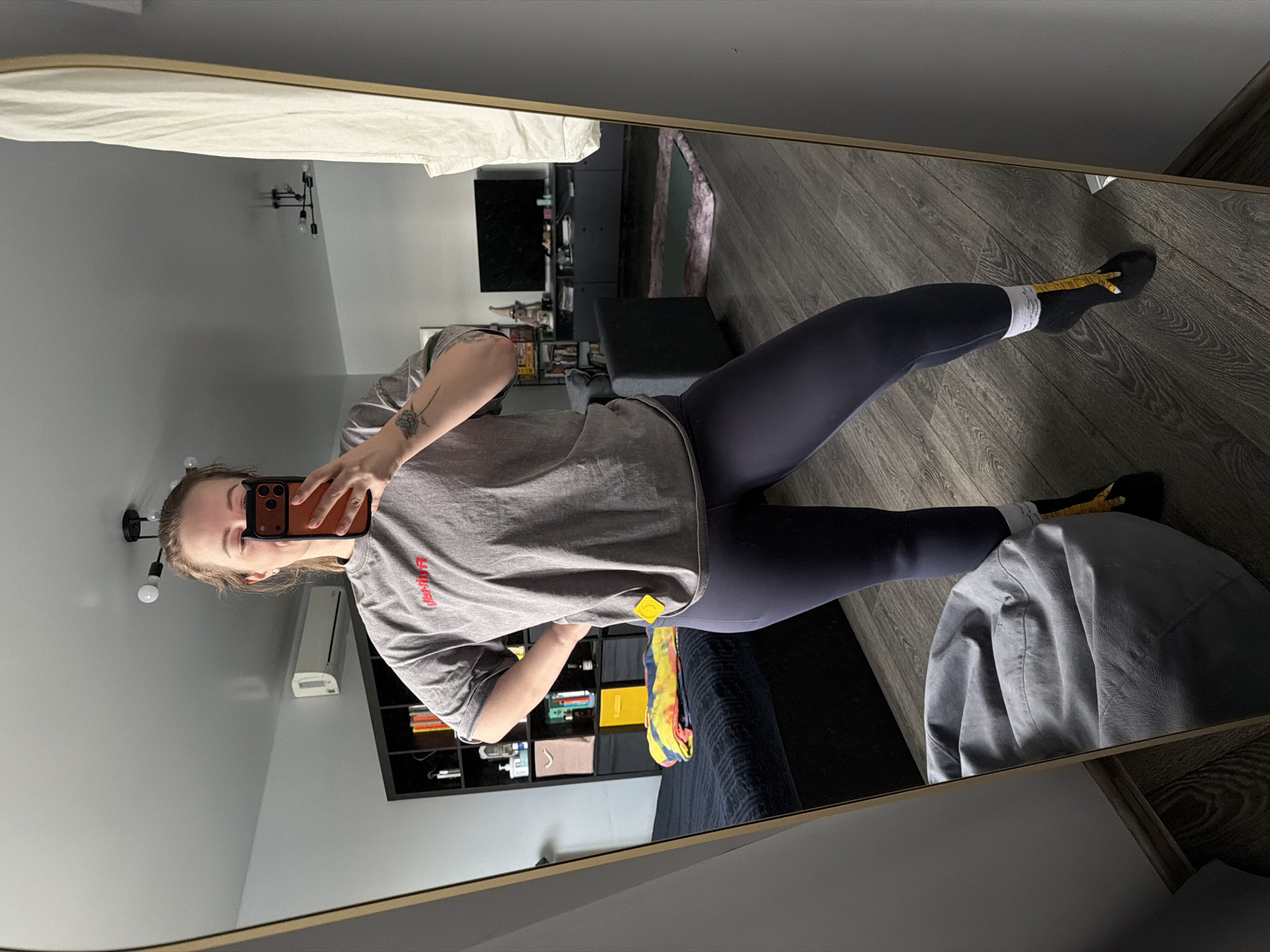 A person is captured in a mirror selfie, standing upright in what appears to be a living room or bedroom. The individual, seen from the waist up, wears a loose-fitting grey t-shirt with red lettering (partially visible as "LIP") and dark blue leggings. They are holding a phone with an orange-brown case up to take the photo, partially obscuring their face. A tattoo is visible on their left forearm. They wear black socks, with one foot also showing a yellow measuring tape resting on the floor.
The room reflected in the full-length, gold-framed mirror features light grey walls and dark wood laminate flooring. In the background, there's a dark media console with a television screen. To the left, a dark shelving unit holds books and various objects. A bed with dark blue bedding and a colorful blanket is also visible. Above the shelving unit, a white air conditioning unit is mounted on the wall, and a modern light fixture with exposed bulbs hangs from the ceiling. A grey bean bag chair is seen near the bottom right of the frame. The overall impression is a casual indoor setting during what appears to be daytime, with indoor lighting also active.