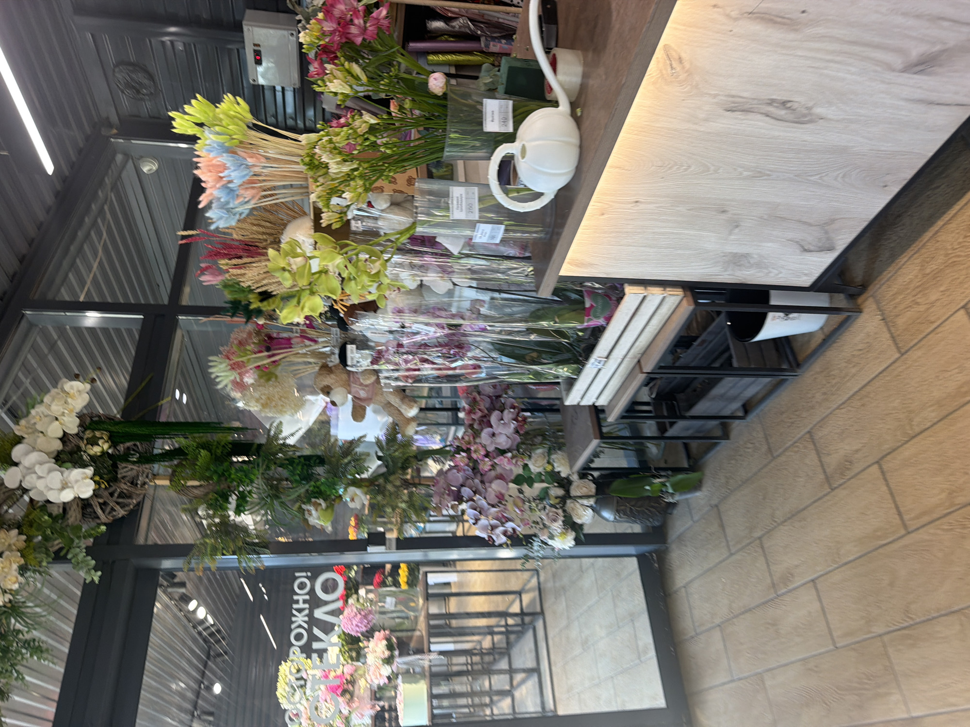 This is a photograph of the interior of a flower shop, likely in Chaikovsky, Russia, given the text visible on the window. The shop features a wide variety of floral arrangements, both fresh and dried, displayed throughout the space. In the foreground, a wooden counter is visible, with a white watering can and some floral supplies on its surface. Behind the counter, an assortment of flowers in glass vases are arranged. To the left of the counter, a large display of orchids and other flowers is prominently featured. The shop's interior is well-lit, with fluorescent lights visible on the ceiling. The flooring is tiled in a wood-grain pattern. The overall impression is one of a bustling and well-stocked flower shop.