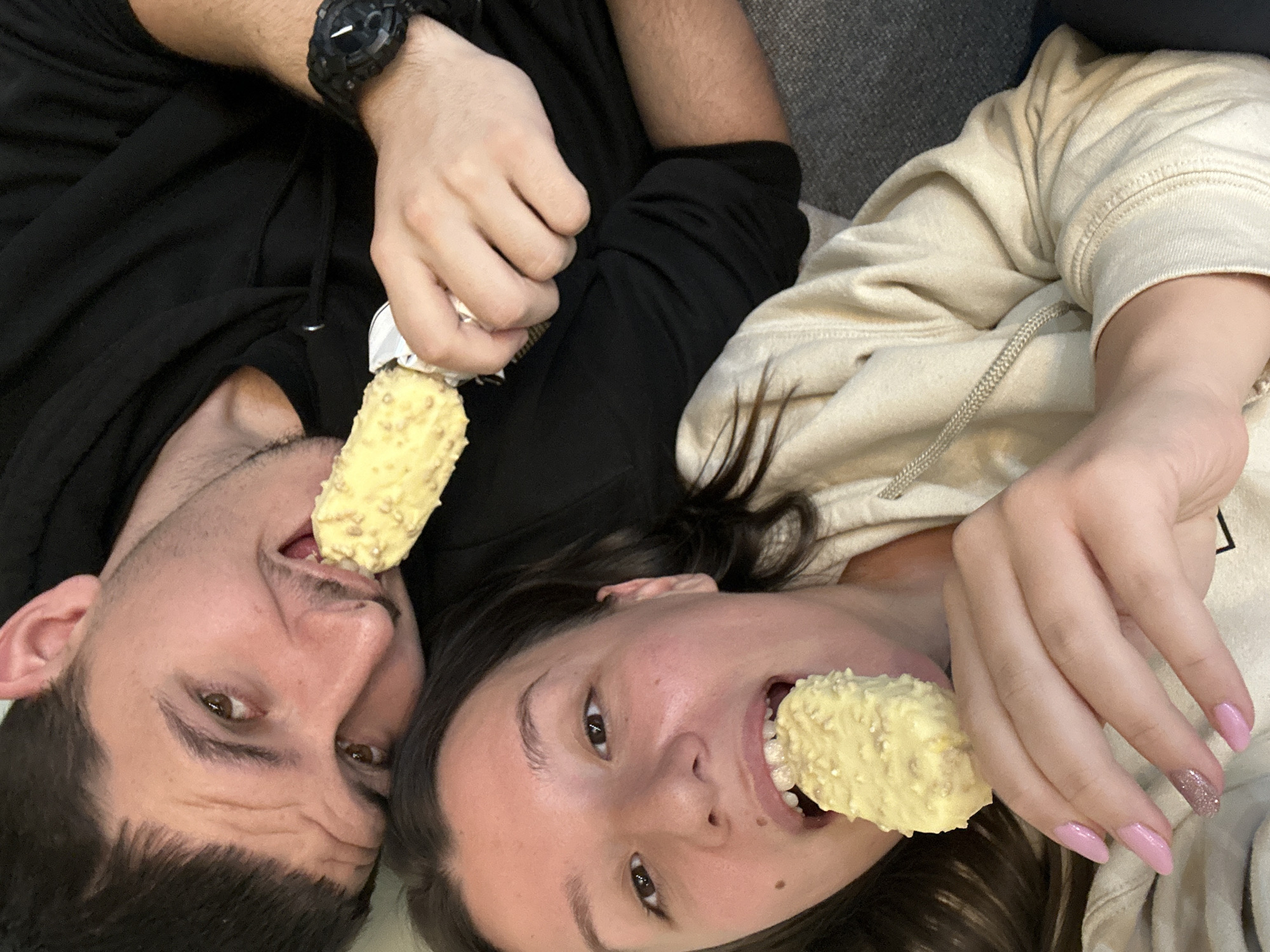 The image displays two individuals, one female and one male, in a close-up, overhead perspective, both consuming identical yellow, almond-coated ice cream bars on sticks. The female is in the foreground, wearing a light-colored hooded sweatshirt. She has dark hair and brown eyes, looking directly at the camera while biting into her ice cream bar. Her right hand, exhibiting pink manicured nails with glitter accent on the ring finger, holds the ice cream. Behind her, the male is partially visible, wearing a black hooded sweatshirt and a dark digital watch on his left wrist. He has short dark hair and stubble, also looking towards the camera with his mouth open around his ice cream bar. The background is out of focus, revealing dark gray upholstered furniture, indicating an indoor environment. No specific location within the city of Sofia, Bulgaria, is discernible from the visual evidence presented.