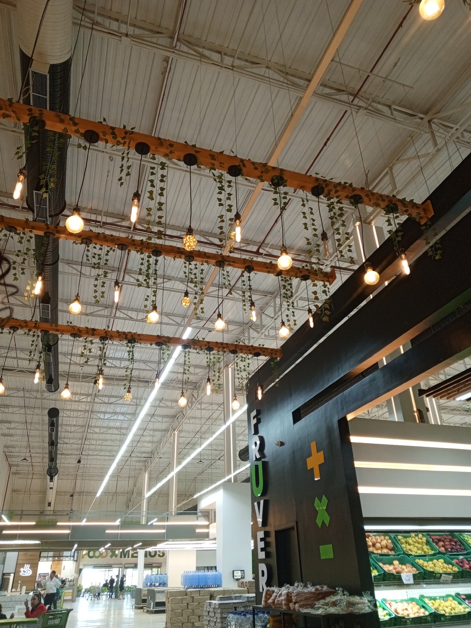 The image shows the interior of a supermarket in Porlamar, Venezuela. The high, white ceiling is supported by metal beams and features industrial air conditioning ducts and two rows of rustic-style wooden beams adorned with hanging light bulbs and artificial vines. In the foreground is a black sign reading "FRUVER" with colorful math symbols, indicating the fruit and vegetable section. Below the sign are shelves displaying various fruits. In the background, shoppers are visible near the "FOOD X METRO" section. Pallets of stacked goods are located in the aisle near the fruit section. The overall lighting is bright and functional.