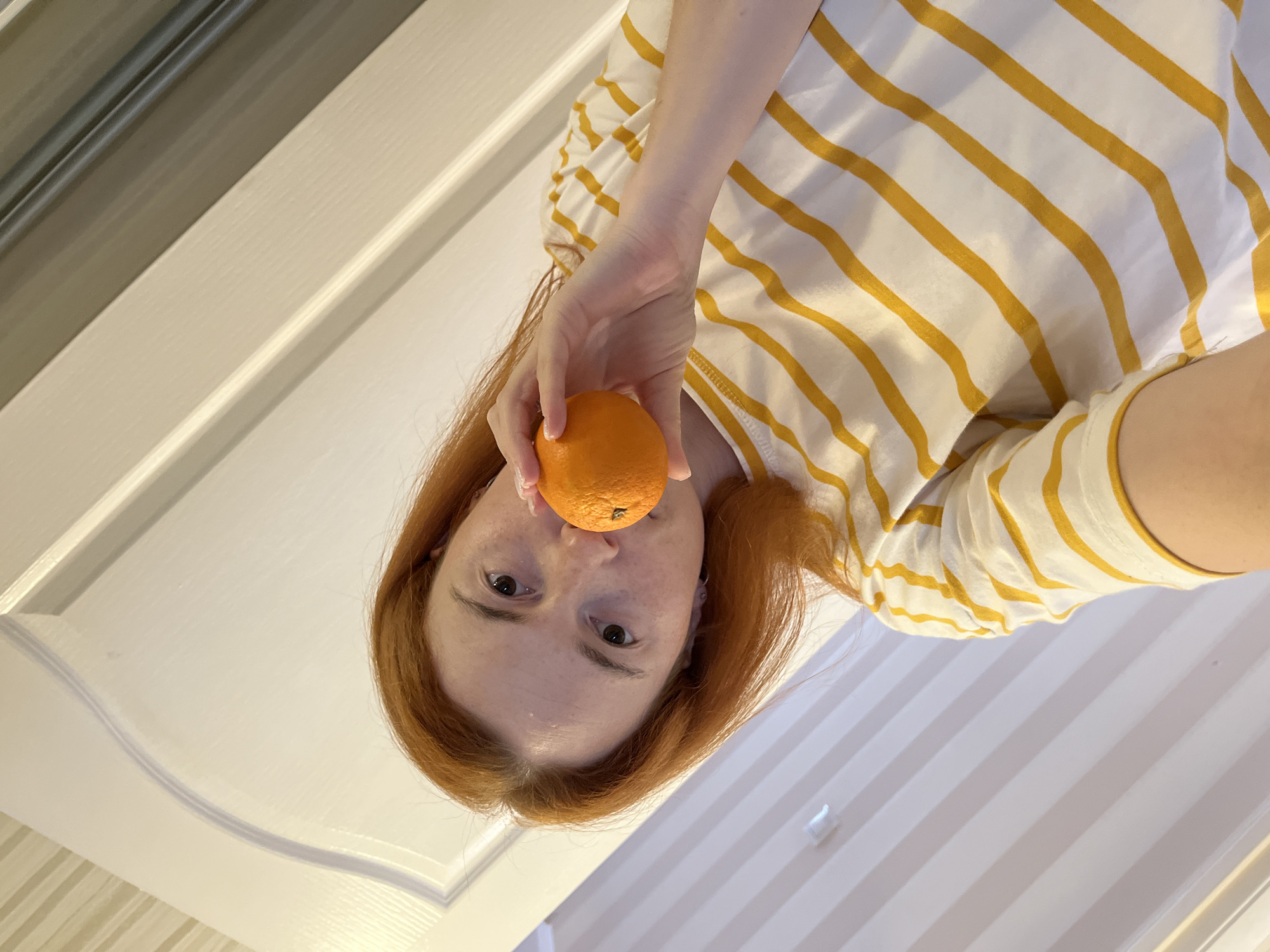 A young woman with reddish-brown hair, wearing a white and yellow striped t-shirt, is holding an orange to her mouth, obscuring her lips. She is looking directly at the camera. The background appears to be an interior setting, possibly a home, with a white paneled door and a wall with vertical pale stripes. The image is taken from a low angle, looking up at the woman. No specific location in Chaikovsky, Russia is identifiable from the visual information provided.