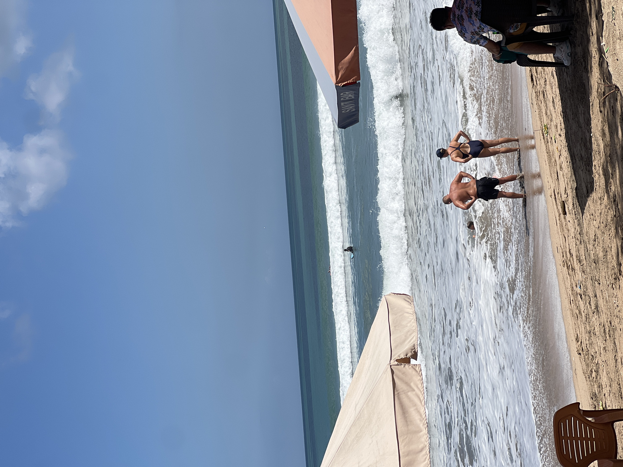 In Kuta, Indonesia, a clear blue sky stretches above a tranquil beach scene. Two people, a man and a woman, stand at the water's edge, their backs to the camera, looking out at the ocean. The man is wearing black swim trunks, and the woman is in a dark blue swimsuit. A man in a floral shirt sits in a dark chair on the right side of the frame, facing away from the water. Further out, a surfer on a blue board rides a wave, and another surfer is visible in the distance. The ocean is calm, with gentle waves rolling onto the sandy shore. Two large beach umbrellas, one tan and one with orange and grey sections, provide shade on the beach. The sand is a light brown color, with some scattered debris. The overall atmosphere is relaxed and peaceful, characteristic of a day at the beach.