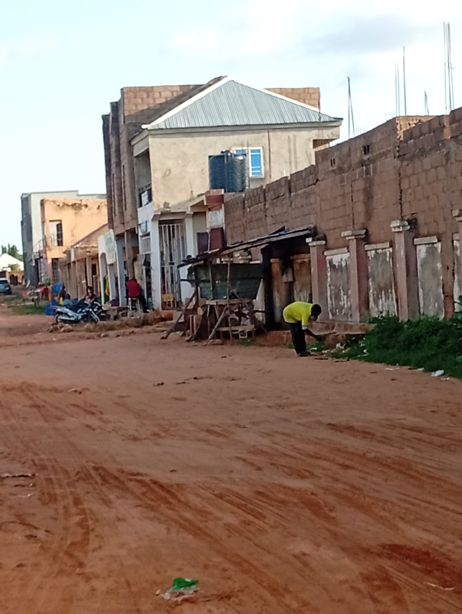 The image depicts a street scene in Tumfure, Nigeria. A dirt road, marked with tire tracks, runs diagonally across the foreground. To the right, a person wearing a bright yellow shirt and dark pants is bending down, appearing to pick up trash from the ground near a low wall constructed of reddish-brown bricks. Beyond the wall, a row of buildings lines the street. One building on the left is a light-colored, multi-story structure with a prominent metal roof. To its right, another building displays a brick facade and a series of doorways or windows. Further down the street, a white motorcycle is parked. People are visible near the motorcycle and in front of some of the buildings, though their features are not clearly discernible. The sky is overcast. The overall impression is of a developing or somewhat unkempt urban environment.