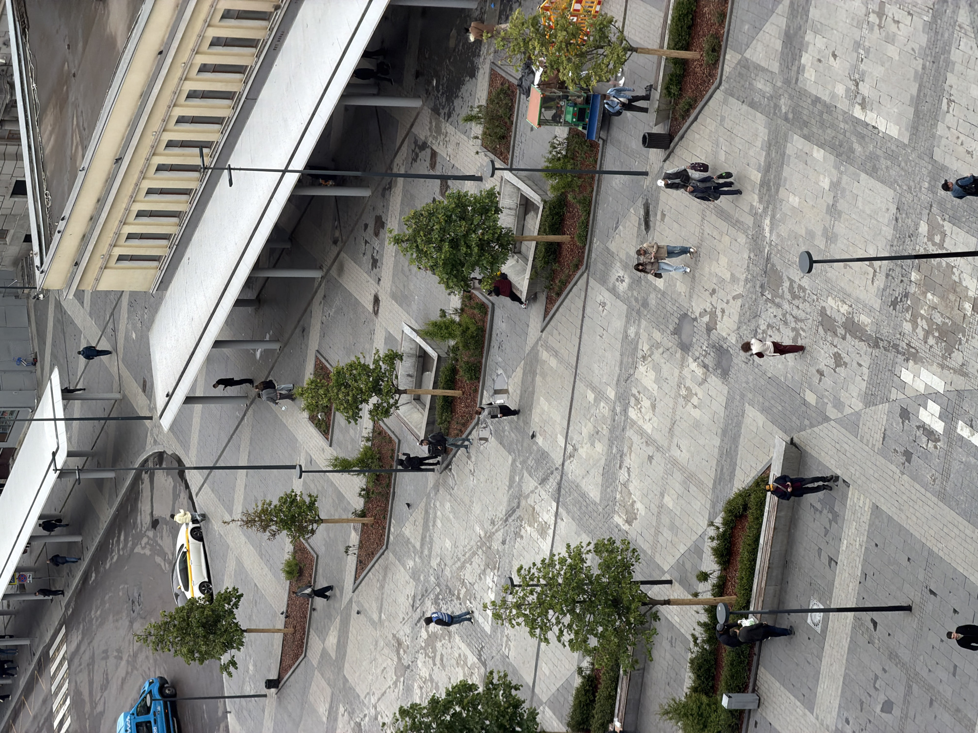 The image depicts an aerial view of an urban plaza in Moscow, Russia. The plaza features a patterned pavement of gray tiles and is bordered by a building with a white facade and a series of repeating rectangular openings. Rows of young trees in raised planters with mulch are spaced throughout the plaza, along with benches and modern light poles.
Several individuals are scattered across the plaza, some walking alone or in pairs, others standing or sitting on benches. A yellow and white taxi is parked near the building, and a blue delivery truck is parked nearby. The scene appears to be a public space used by pedestrians.