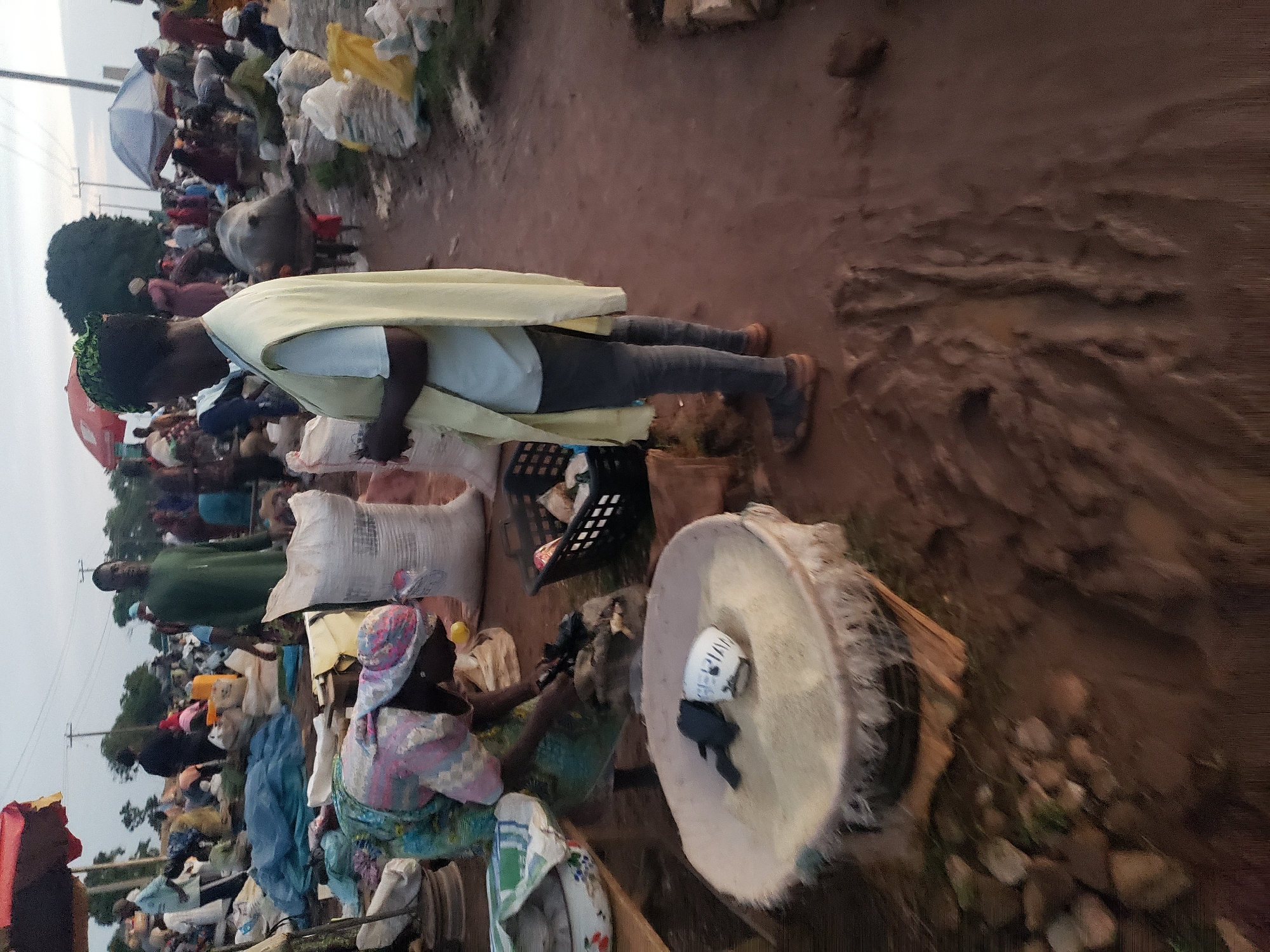 The image depicts a bustling outdoor market scene in Kakuri Gwari, Nigeria. A woman, dressed in a colorful patterned headwrap and traditional attire, is bent over a large, shallow bowl filled with a light-colored grain, likely rice. She is actively scooping or measuring something from a sack with her hands. To her right, a woman in a pale yellow vest and dark pants stands, looking to the side. Behind her, a few large white sacks are visible, along with a black basket containing various items. In the background, the market is crowded with people and stalls, indicated by numerous figures, tents, and piles of goods, including more sacks. The ground is a mix of dirt and mud, suggesting a recent rain or general market conditions. The overall atmosphere is that of a busy marketplace, likely in the late afternoon or early evening due to the lighting.