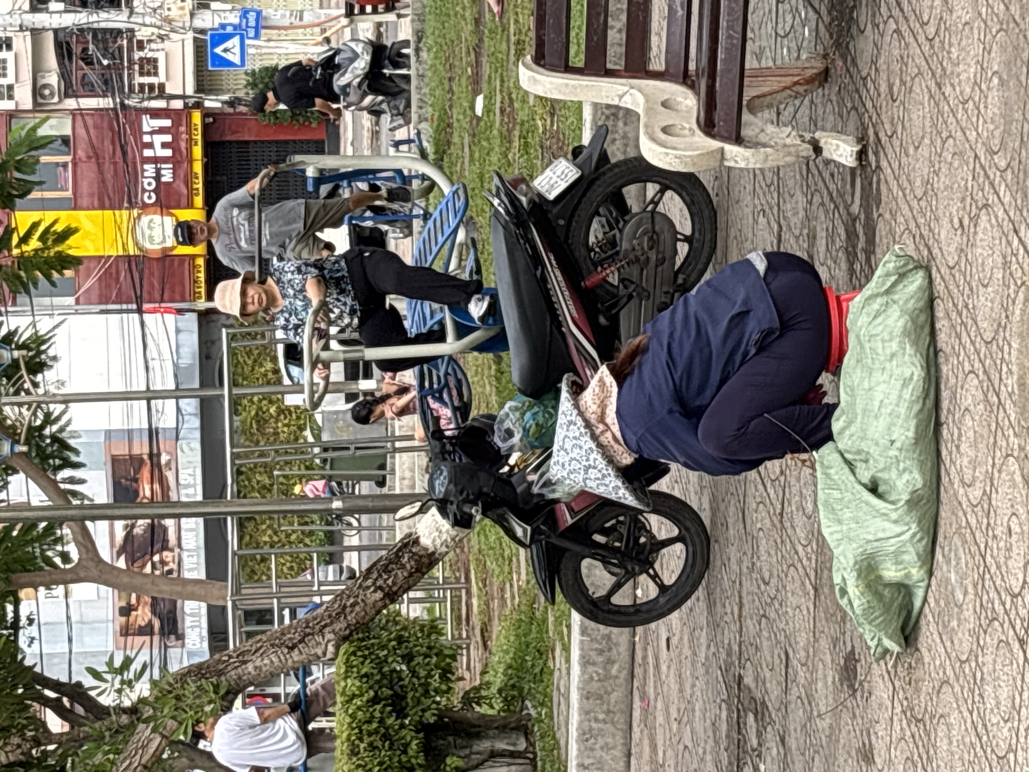 The image depicts a street scene in Nha Trang, Vietnam, likely in a park or public square. In the foreground, a person is crouched down beside a large, light green tarp on the ground. They are wearing a dark blue jacket, dark blue pants, and a patterned conical hat, and are sitting on a small red stool. A black motorcycle is parked behind them, with a green bag on its seat.
In the middle ground, two people are using outdoor exercise equipment. A woman wearing a light-colored hat and a black and white patterned top is seated on a machine, with her legs extended. A man in a grey t-shirt and dark shorts stands behind her, holding onto the equipment. Behind them, a person in a black shirt walks away from the camera.
The background shows buildings, including one with a red and yellow facade and signage that reads "CƠM MÌ HT". A blue traffic sign with a white triangle and a pedestrian symbol is visible. Trees and other greenery are present, as is a metal fence. The ground is paved with patterned tiles. The scene appears to be an outdoor public space during the day.