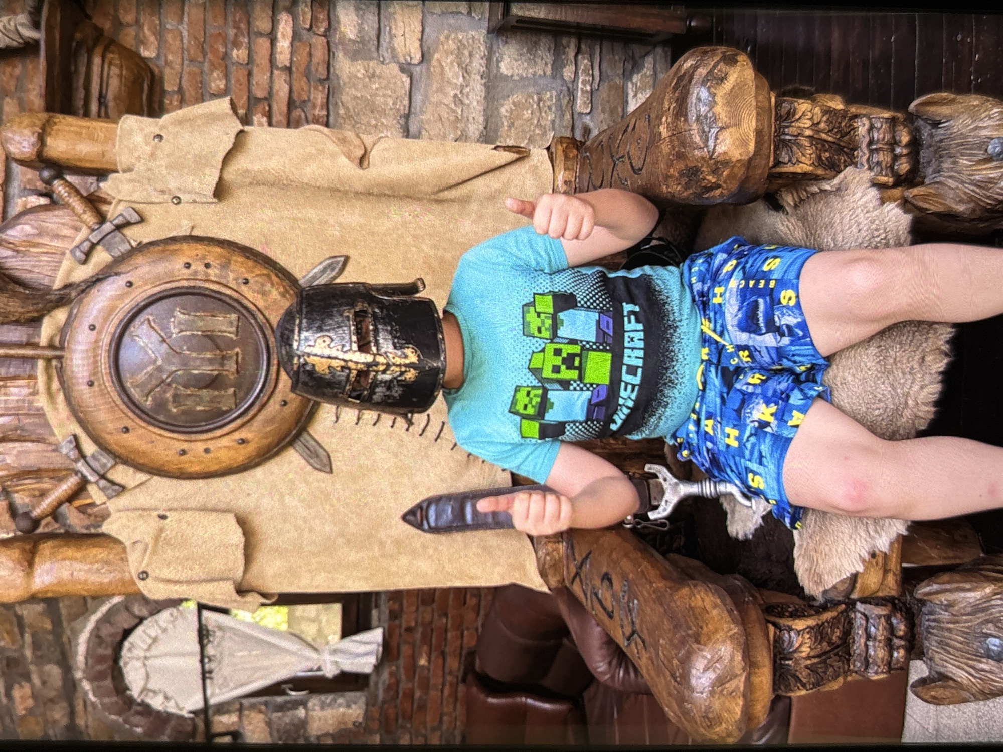 An indoor, vertically oriented image captures a child posing enthusiastically with a large, elaborate wooden figure, likely a chair or statue, adorned with medieval-themed props. The scene is set against a rustic background of brick and stone walls, with some wooden architectural elements.
The child, seen from the waist up, wears a dark, metallic-looking helmet resembling a knight's visor, partially obscuring their face. They are dressed in a light blue t-shirt featuring pixelated characters and the prominent text "MINECRAFT". Their blue shorts have a patterned design with yellow letters, including the visible word "BEACH". The child's left hand is giving a thumbs-up, while their right hand holds a dark, metallic object that appears to be a stylized weapon or tool.
The large wooden figure, crafted from carved wood, serves as the main prop. It features substantial wooden arms and legs, with detailed carvings and accents of light brown fur. Draped over the figure is a tan, leather-like fabric. Attached to this is a round, wooden shield with metallic studs and a central emblem. Several small, metallic-looking swords or daggers are visible around the figure and shield, completing the medieval or fantasy aesthetic. The overall impression is one of playful engagement with themed decor.