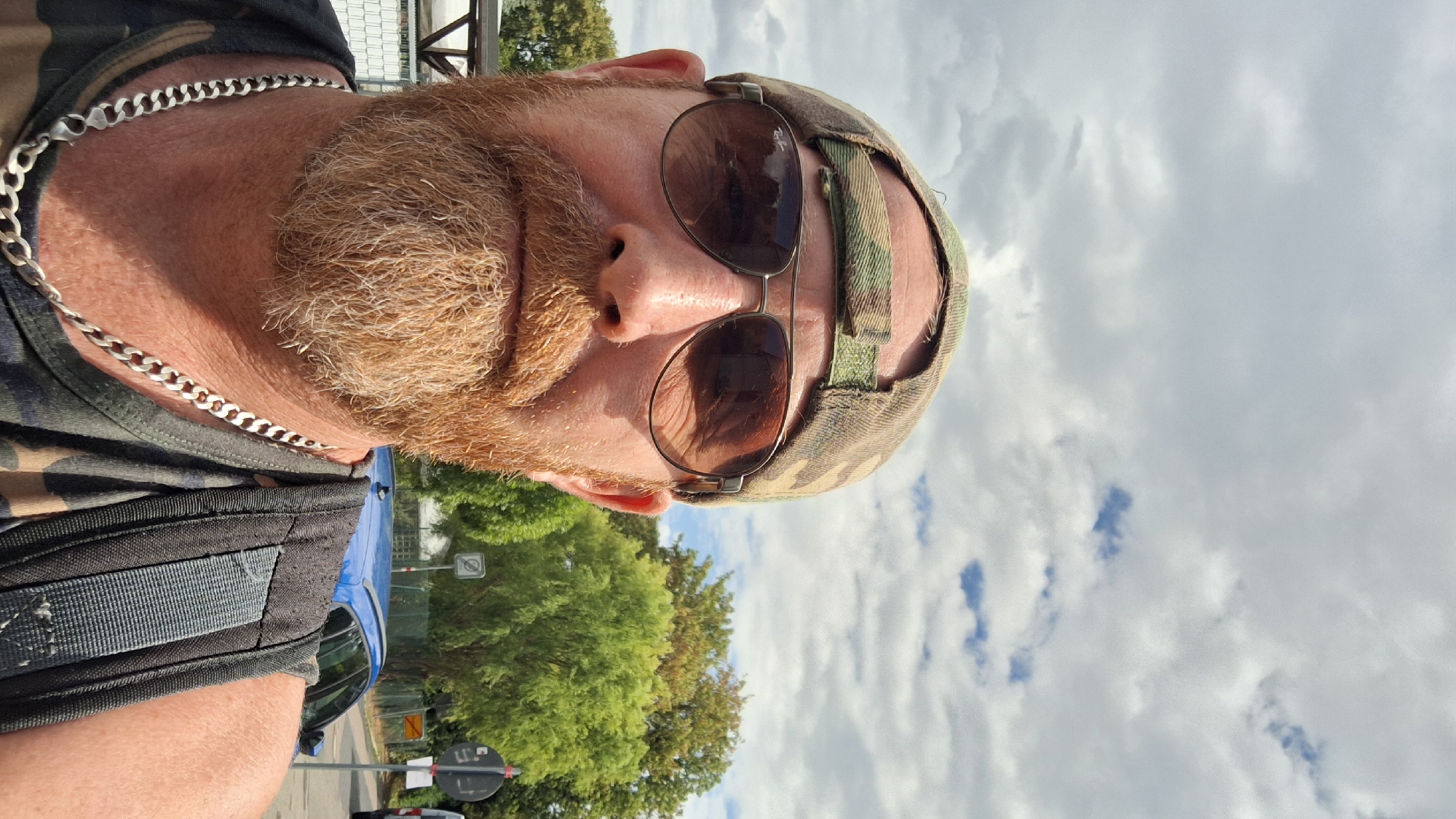The image is a selfie of a man wearing a camouflage baseball cap and sunglasses. He has a red beard and is wearing a silver chain and a camouflage tank top. A backpack is visible on his right shoulder. The background shows trees, a blue car, and a cloudy sky. There are some street signs and a fence. The location is identified as being in Karlsruhe, Germany.