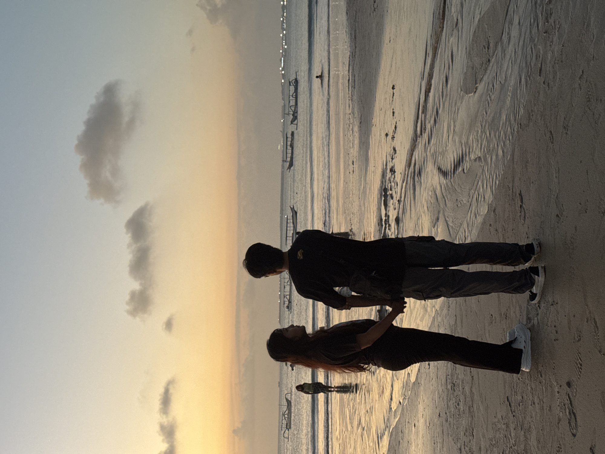 In Kuta Selatan, Indonesia, a couple stands hand-in-hand on a sandy beach during sunset. The man, on the right, wears a black t-shirt and gray cargo pants, with a dark bag slung over his shoulder. The woman, on the left, is dressed in a dark t-shirt and dark pants. Their backs are to the camera as they face the ocean. In the background, several traditional Balinese fishing boats, known as jukung, are anchored in the calm water. A lone figure is swimming in the sea further out. The sky displays a gradient of pale blue and warm orange hues, with scattered clouds. The wet sand reflects the fading light, creating shimmering patterns. The overall scene captures a serene moment at the end of the day by the ocean.