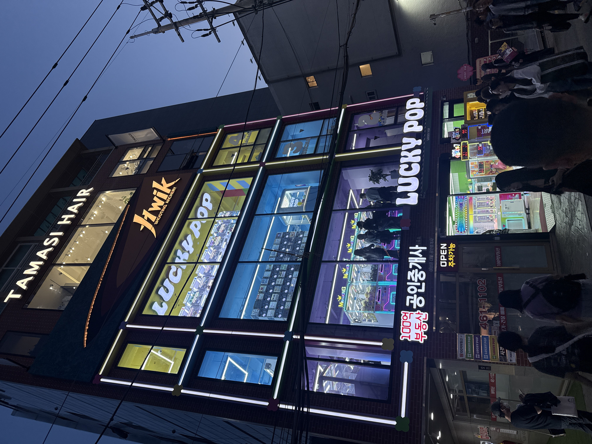The image shows a brightly lit, multi-story building at dusk in Seoul, South Korea. The building's facade is a brick and glass grid, accented with neon lights. The building features signs for "TAMASI HAIR" on the top floor, "JHwik SHOOTING AFICHERY" below that, and "LUCKY POP" prominently displayed in large neon letters spanning several floors. Below is an office of estate agents. The windows display arcade claw games and other products. People are gathered on the sidewalk in front of the building. Electrical wires stretch above the building. The lighting is a combination of the building's neon and the twilight sky.