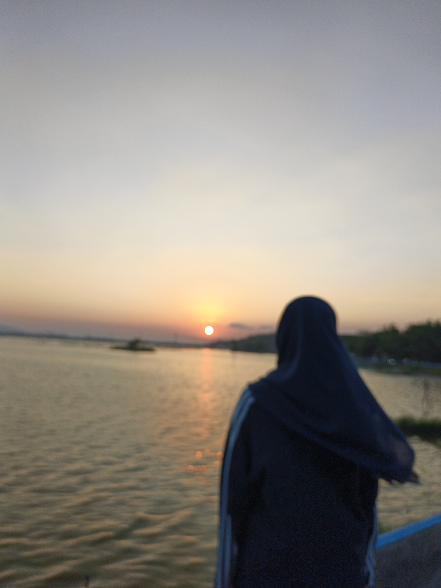 An over-the-shoulder, slightly blurred medium shot shows a person wearing a dark blue hijab and jacket standing on a walkway, facing a body of water at sunset. The sun is visible as a bright orb on the horizon, casting a warm glow on the water, which has a rippling texture. In the distance, a faint outline of trees and land is visible. The sky is a soft gradient of pale blue and orange. The person appears to be observing the sunset over the water. The location is identified as Krakitan, Indonesia.
