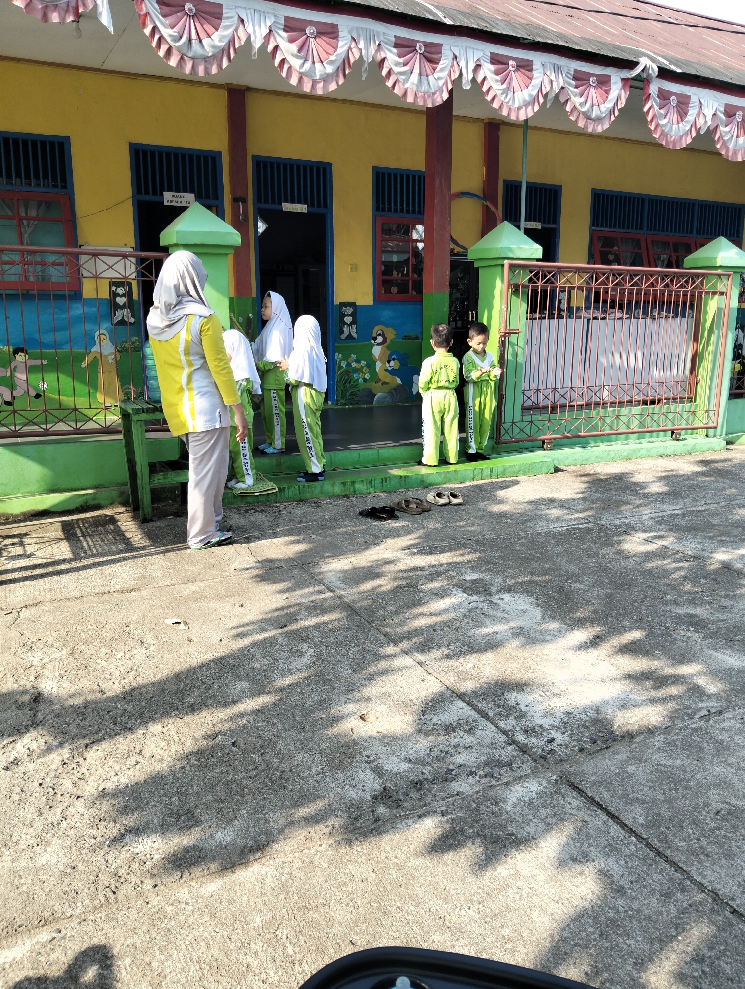 The image captures an outdoor scene in front of a brightly colored building, possibly a school, in Janju, Indonesia. The building is painted yellow with blue window frames and red accents around some windows. A decorative banner with red and white swags hangs across the top of the building. A green fence runs along the front of the property, with a metal gate. On the wall of the building, a mural depicts cartoon-like figures.

Several people are present. In the foreground on the left, a woman wearing a grey hijab and a yellow and white long-sleeved top and light grey pants stands facing away from the camera. To her right, three children in matching green uniforms and white hijabs are standing on a raised platform in front of the building. Two of the children appear to be young boys, and one is slightly behind them. To the far right, another child in a similar green uniform and white hijab stands by the fence.

On the concrete ground in front of the building, several pairs of sandals are neatly placed. The ground is paved with concrete slabs, and dappled sunlight creates shadows from unseen trees. The overall impression is a sunny day in front of an educational facility, likely a kindergarten or primary school, given the colorful building and the young children present.