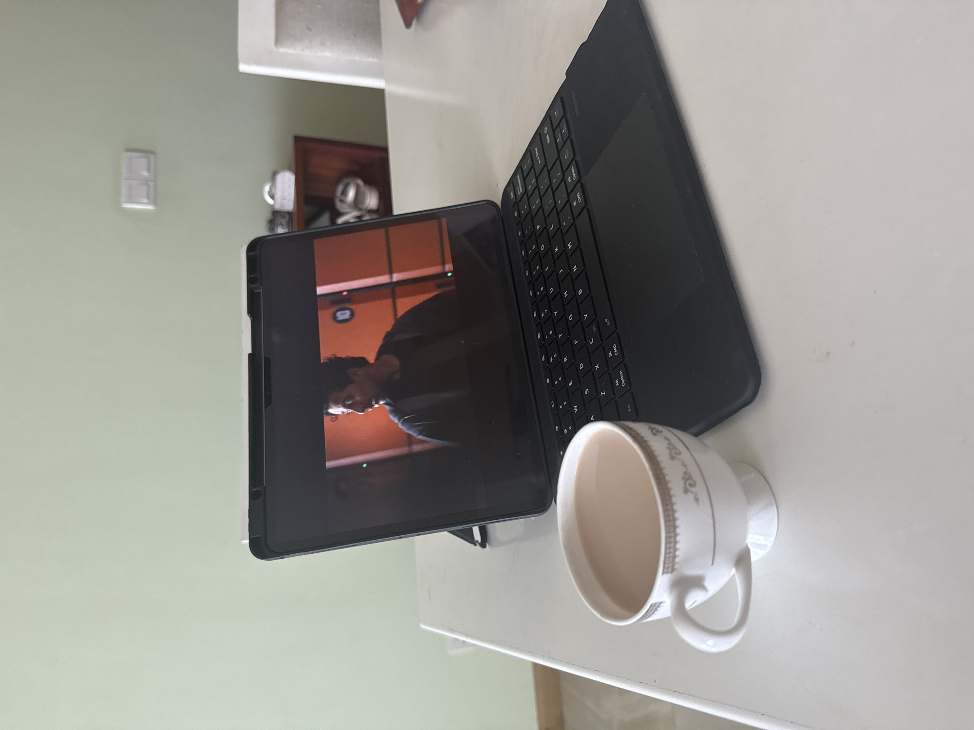 A tablet device with a keyboard is placed on a white table. The tablet screen displays a scene from a movie, showing a person. A white teacup with a decorative rim is positioned to the left of the tablet, filled with a light brown beverage. The background features a light green wall with two light switches. In the far background, a wooden cabinet with some items on top is visible. The scene is set indoors, likely in a residential setting, possibly in Khan Mean Chey, Cambodia.