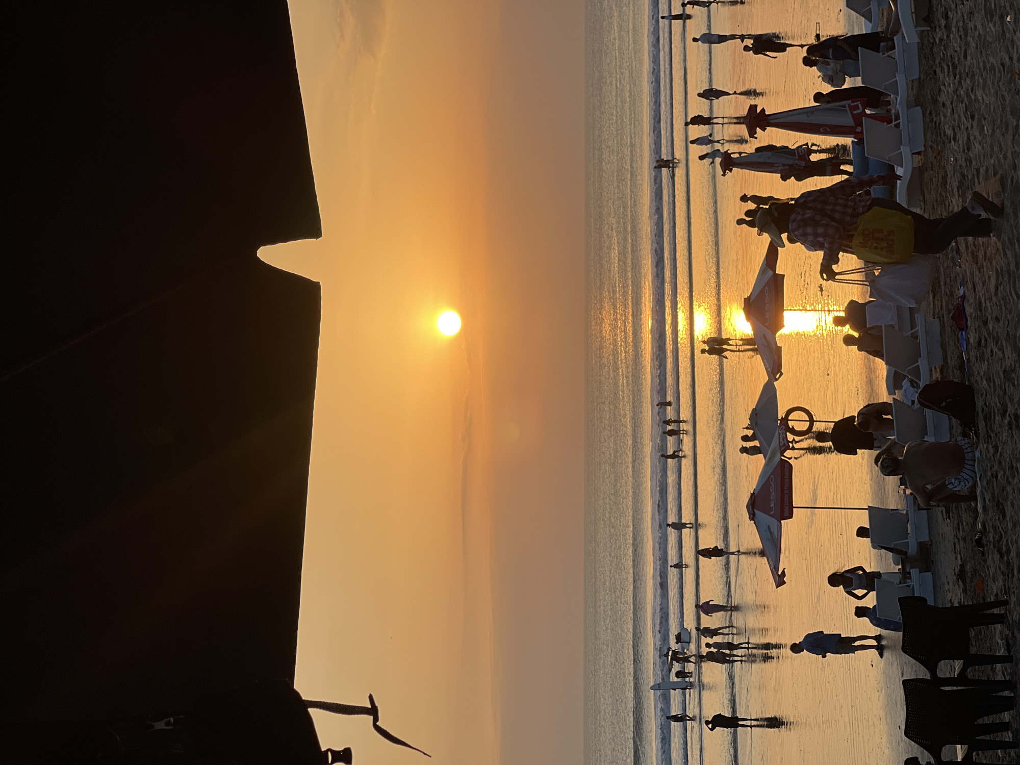 The image depicts a crowded beach scene at sunset in Kuta, Indonesia. The sun, a brilliant orange orb, is low on the horizon, casting a warm glow across the water and wet sand. Silhouettes of numerous people are visible, wading in the shallow water, walking along the shoreline, and relaxing on beach chairs under umbrellas. Some individuals are carrying surfboards, suggesting a popular surf spot. The atmosphere appears lively and relaxed, with the setting sun creating a picturesque backdrop. The foreground is partially obscured by a dark, possibly fabric structure, such as a beach umbrella or awning, casting a shadow over the initial part of the scene.