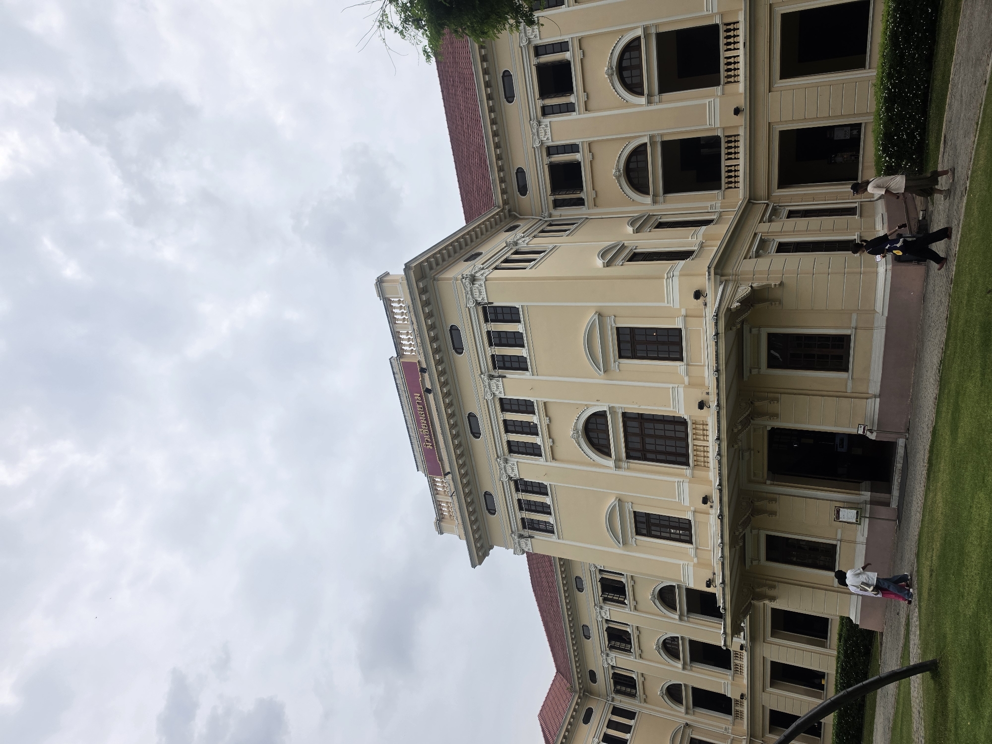 The image depicts a building in Bangkok, Thailand. The structure is a multi-story, yellow-colored building with a red roof and many arched windows. The building has decorative molding. The sky is overcast, and a tree branch is visible near the top right corner. People are walking along the sidewalk adjacent to the building. Green grass and bushes border the sidewalk.