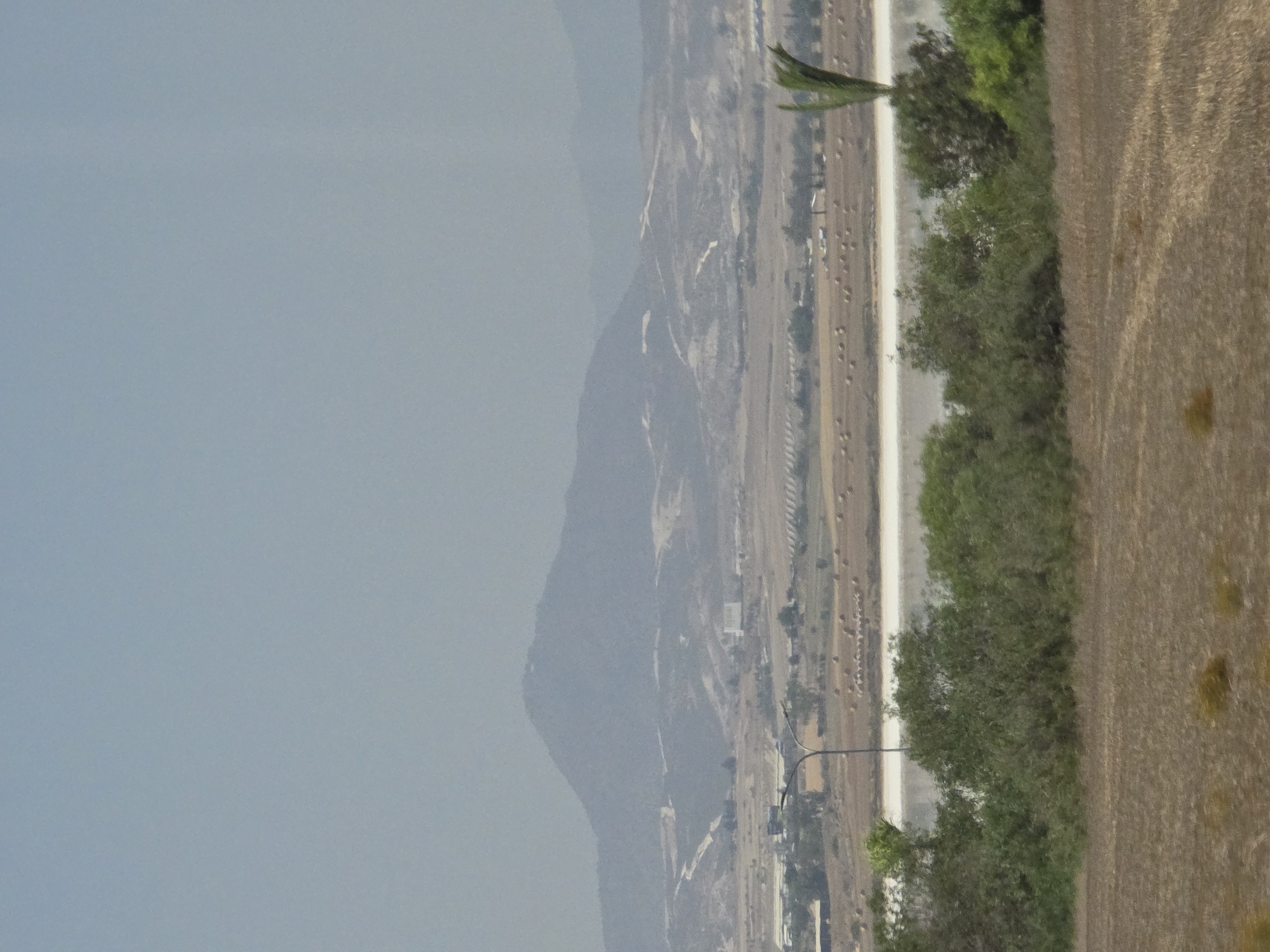 The image shows a panoramic view of a landscape near Larnaca, Cyprus, likely encompassing the area around the Larnaca Salt Lake. In the foreground, a dry, cultivated field with sparse, yellowish-brown vegetation is visible. In the mid-ground, a body of water, possibly the salt lake, appears as a bright, reflective band. It is bordered by lush green trees and bushes. A modern street light with a curved pole stands to the left of the water. Beyond the water, the landscape transitions into a dry, arid terrain with scattered fields of hay bales. In the background, a large, hazy mountain range dominates the horizon. The sky is a pale, overcast blue, with a light haze obscuring distant details. The overall scene suggests a rural or semi-rural environment under a subdued, possibly dusty, atmospheric condition.
