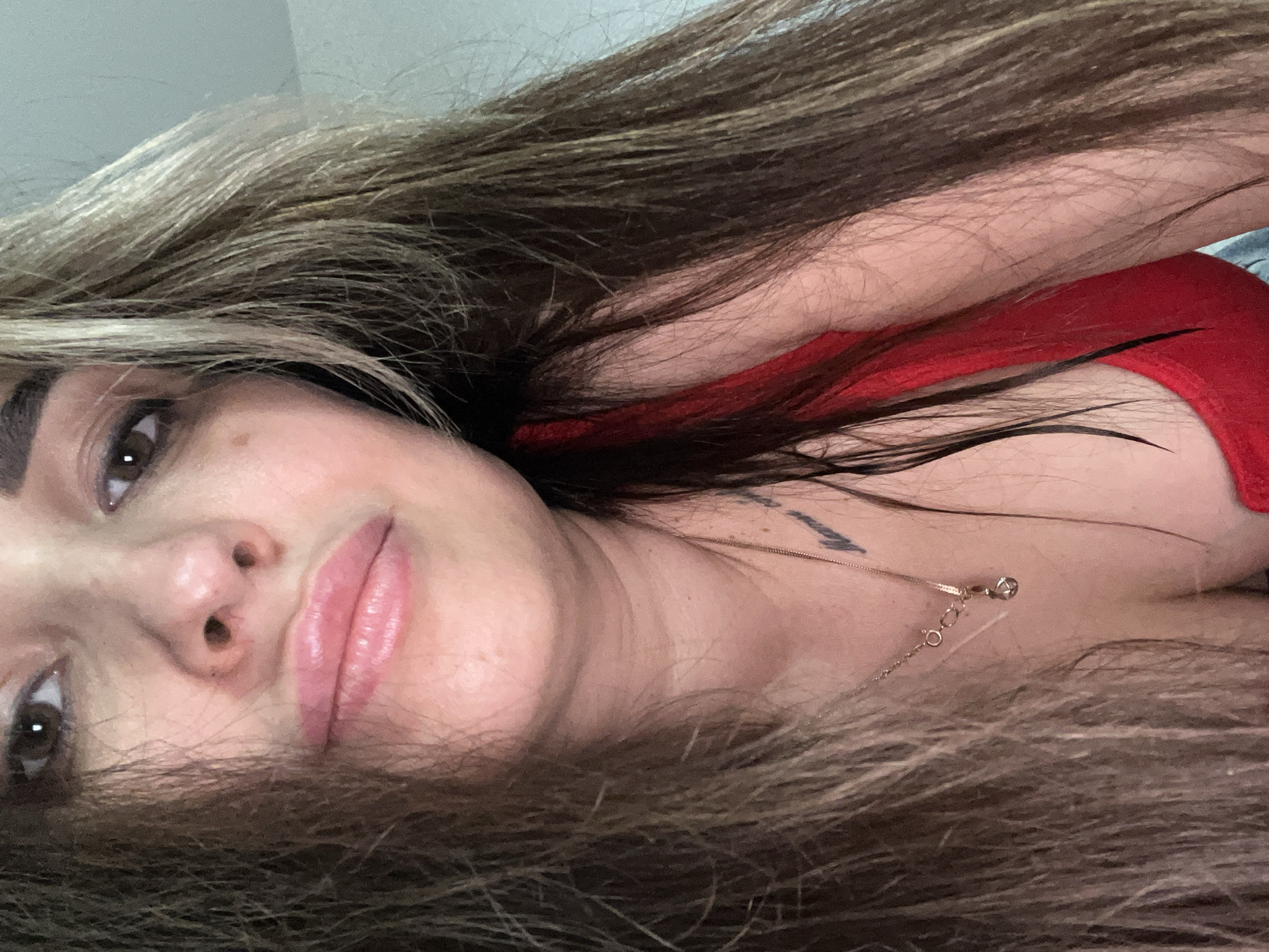 The image is a close-up, selfie-style portrait of a young woman lying down. Her head is positioned on the left side of the frame, tilted to the side, with her body extending to the right. She has long, wavy brown hair with lighter highlights, which is spread around her head and shoulders, framing her face and partially covering her body. Her brown eyes are open and looking directly at the camera, and her full lips are slightly parted, revealing a soft pink color. She has clear skin with a few small moles or freckles visible on her face.
She is wearing a red top, possibly a tank top or camisole. A delicate gold-colored chain necklace with a small, clear, possibly diamond or cubic zirconia, pendant hangs around her neck. A portion of a dark-ink tattoo, which appears to be text or a symbol, is visible on her upper right shoulder or arm. The lighting is soft and even, highlighting her features. The background is largely obscured by her hair and body, with a light-colored surface visible at the top. Her expression appears neutral and calm. There is no visible text in the image.
