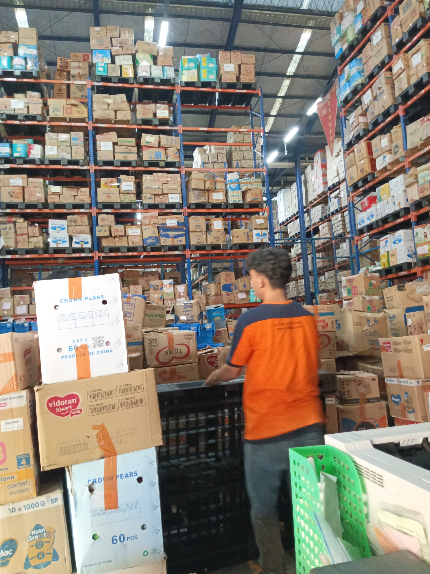 The image shows a warehouse interior filled with shelves stacked high with cardboard boxes of various products. The boxes are diverse in size, color, and labeling, suggesting a range of consumer goods. In the foreground, a young male wearing an orange and dark blue t-shirt and dark pants is standing, facing away from the camera and towards the shelves. He appears to be a worker in the warehouse. To his right, a white office printer and a green plastic organizer containing papers are visible. The warehouse has a high ceiling with exposed industrial lighting and metal support structures. The overall impression is a busy operational environment for storage and distribution. The presence of specific brands and "Produce of China" labels on some boxes indicates that this is likely a distribution center for imported goods. Although no direct visual cues place the warehouse in Makassar, Indonesia, the context of a large distribution center storing various consumer products is consistent with the city's role as a major port and commercial hub in eastern Indonesia.