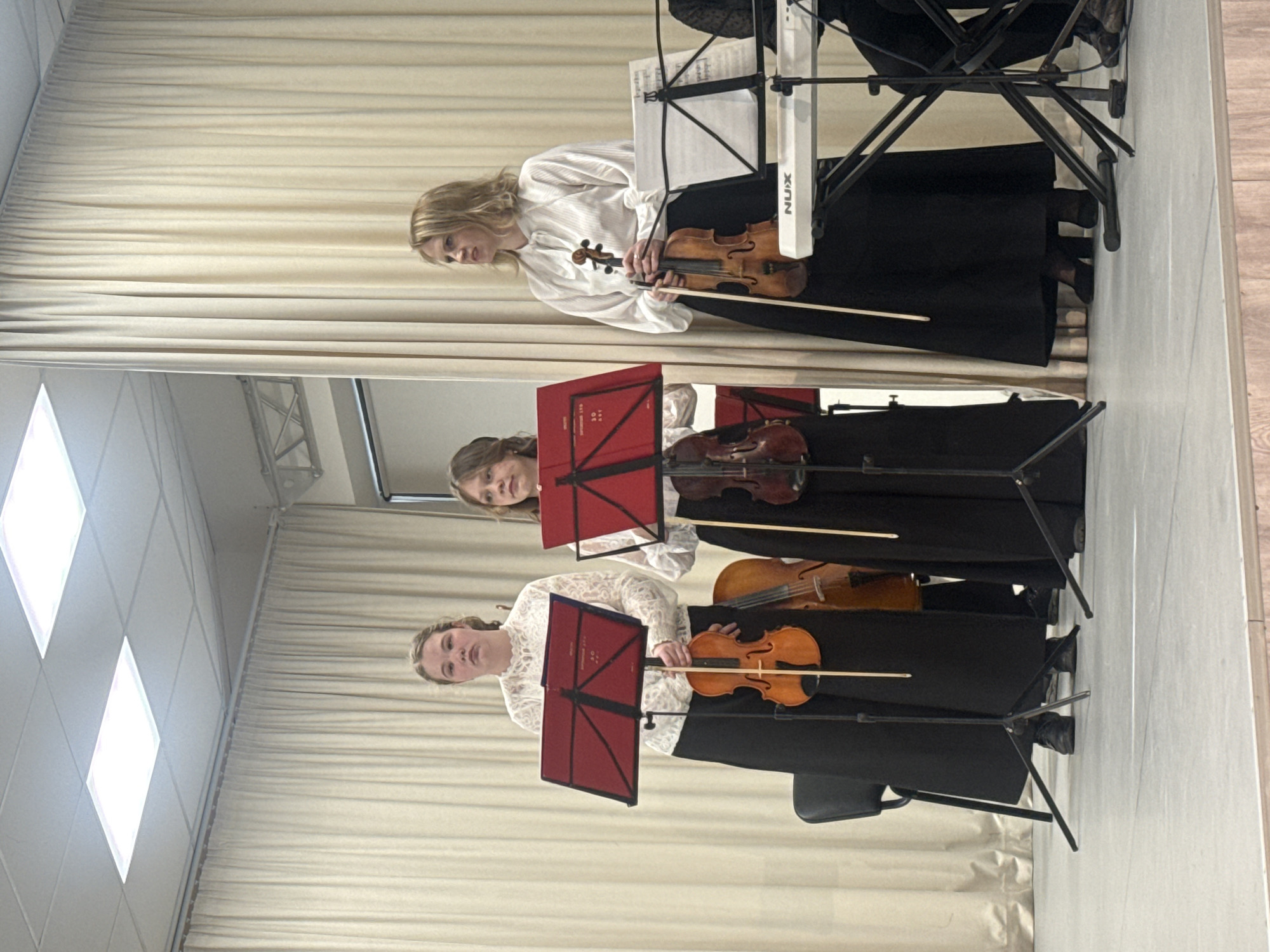 Three women are positioned in front of a beige curtain, each holding a violin and bow. They are dressed in white blouses and long black skirts. Each woman has a red music stand in front of her. The room appears to be indoors, possibly a concert hall or performance space. The location and city are unknown.