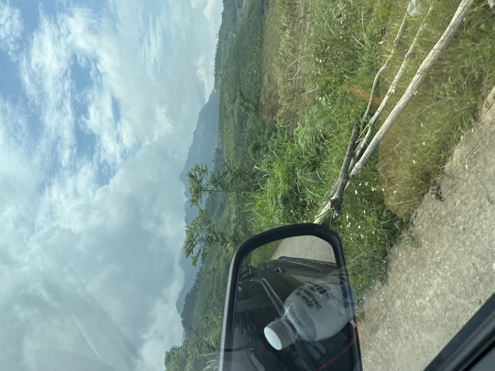 Captured from inside a vehicle, the image displays a mountainous, verdant landscape in Suối Giàng, Vietnam. The sky is partly cloudy with scattered white clouds. The side mirror of the vehicle reflects a water bottle. The foreground includes roadside foliage and a section of a concrete road.