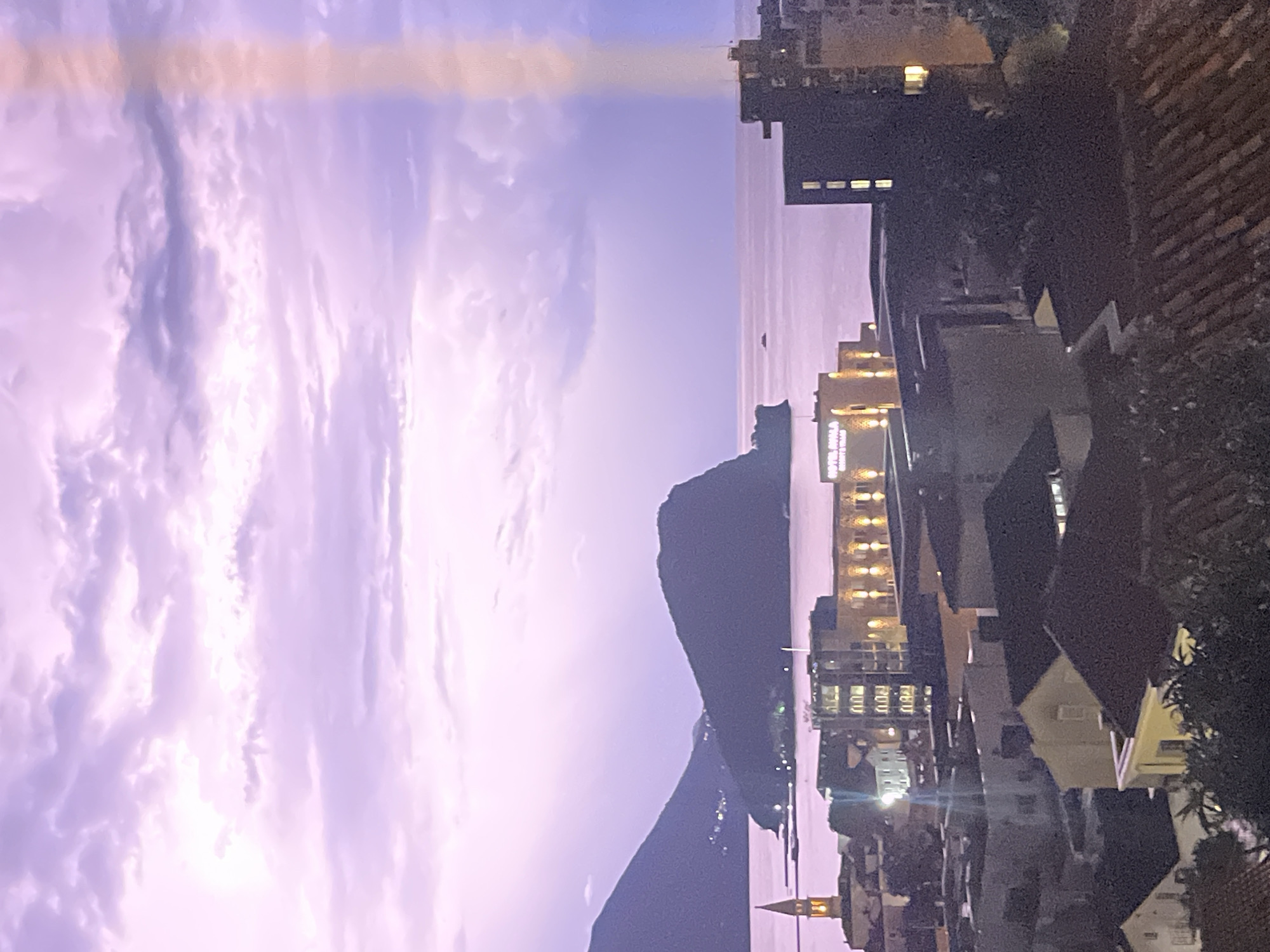 The image captures a twilight view of Budva, Montenegro, characterized by a serene atmosphere. The sky is a gradient of soft purple and pink hues, with scattered clouds suggesting a fading sunset or pre-dawn light. A distinct vertical light streak is visible in the upper right quadrant of the sky, possibly a lens flare or artifact.
In the middle ground, a body of water, likely the Adriatic Sea, stretches towards distant, dark-silhouetted hills. The most prominent of these is a large, rounded island or headland, with a smaller, conical hill positioned behind it and to its left. Lights are scattered across the slopes of these landmasses, indicating settlements or structures.
The foreground and mid-ground are dominated by the cityscape of Budva. Numerous buildings of varying heights and architectural styles are visible. To the far left, a tall, slender church tower with a lit spire stands out. A prominent building with a light-colored facade and illuminated windows reads "HOTEL AVILA RESORT & SPA" in the middle-left of the frame. Adjacent to it are other structures with glowing windows, suggesting occupancy. To the right, darker, more modern buildings with illuminated windows are interspersed with lower-rise structures. The rooftops of several buildings, some with terracotta tiles and others with darker materials, are visible in the lower portion of the image. Trees with dark foliage are also present, partially obscuring some of the buildings. The overall impression is of a coastal city transitioning from day to night, with artificial lights beginning to illuminate the urban landscape against the backdrop of natural scenery. No people are discernible in the image.