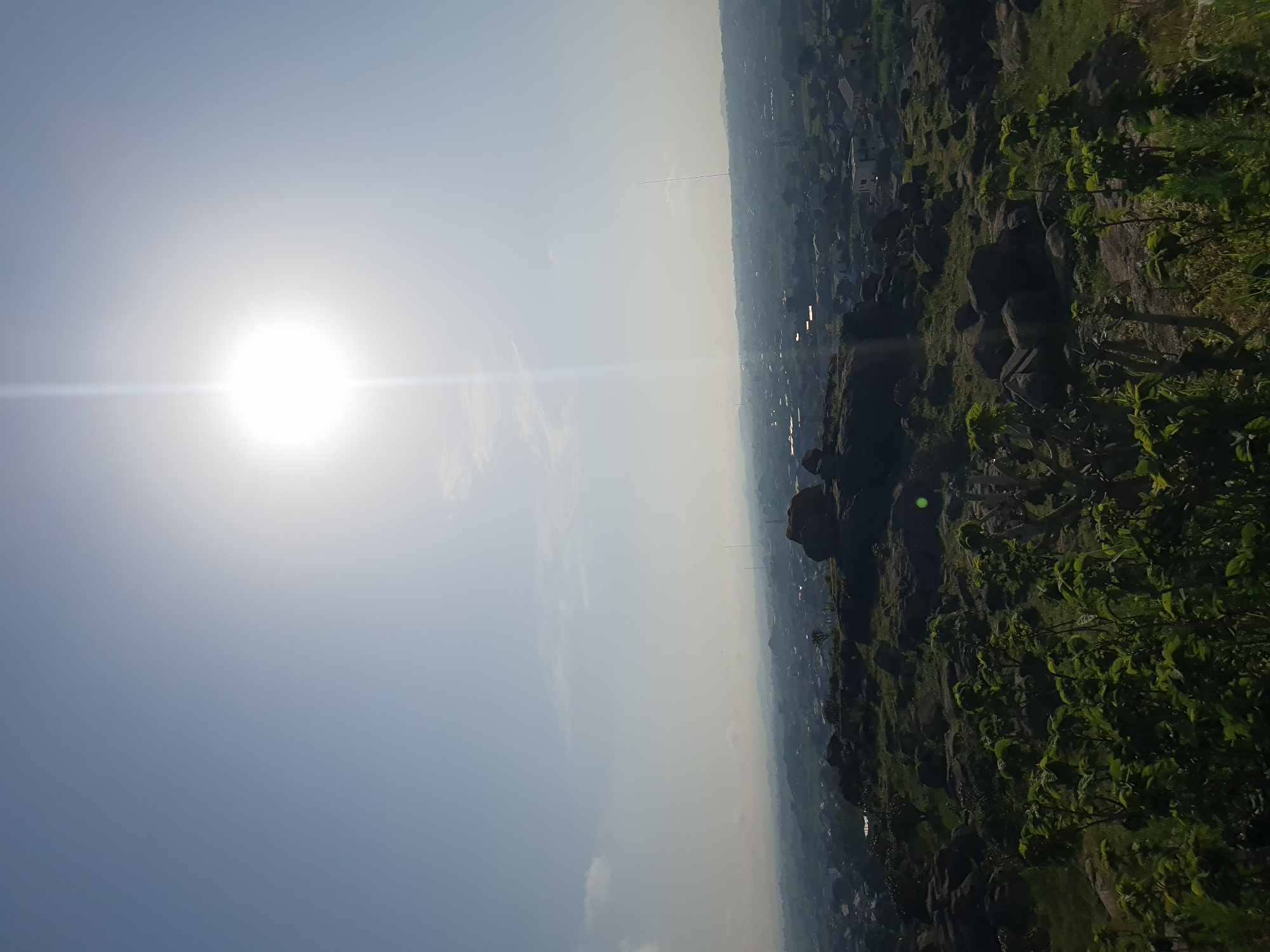 A bright sun shines in the upper part of the frame, creating a lens flare effect. Below, a hazy sky transitions into a panoramic view of a city in the distance. In the foreground, a rocky, grassy terrain is dotted with green foliage. The city appears to be a densely populated area with buildings and structures visible. The overall scene depicts an urban landscape viewed from a higher vantage point, likely Gura Top, Nigeria, under a bright, sunny sky.