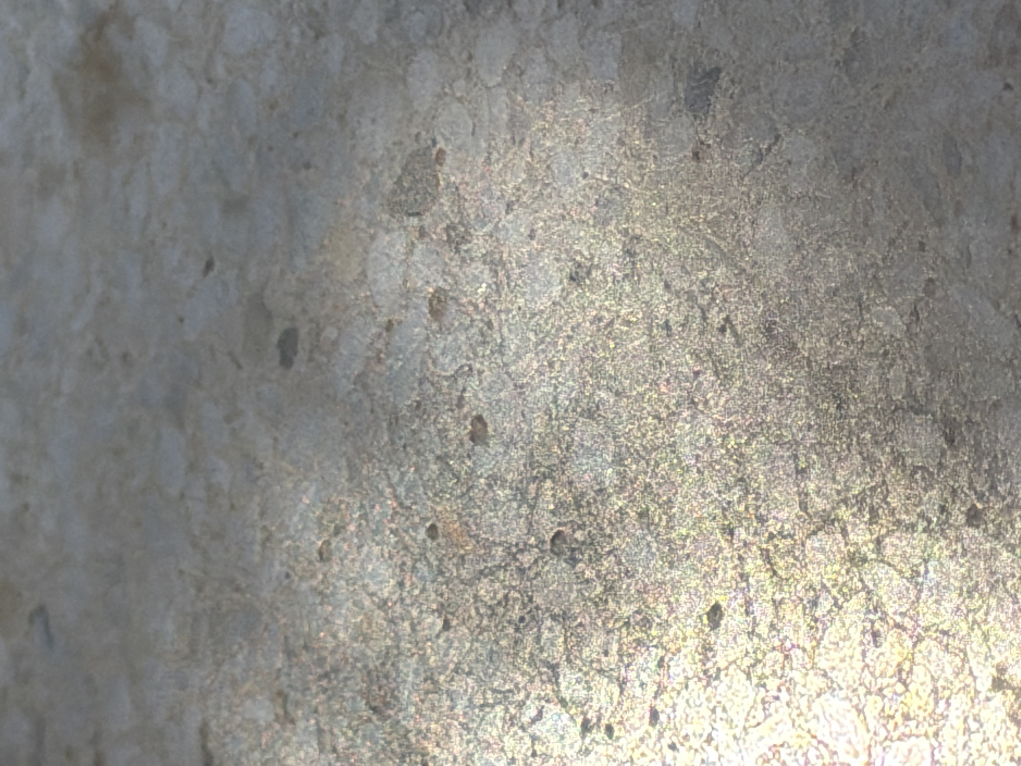 The image displays a close-up, abstract view of a textured surface. It appears to be a stone or concrete material with visible aggregate and some darker inclusions or pores. The lighting creates a play of light and shadow, with a bright, almost iridescent area on the lower left and a more subdued, shadowed area on the upper right. The texture is granular and uneven. There are no discernible people, animals, or recognizable objects in the frame, nor any clear indication of a specific location within Arcahueja, Spain. The focus is entirely on the surface's material properties and the way light interacts with it.