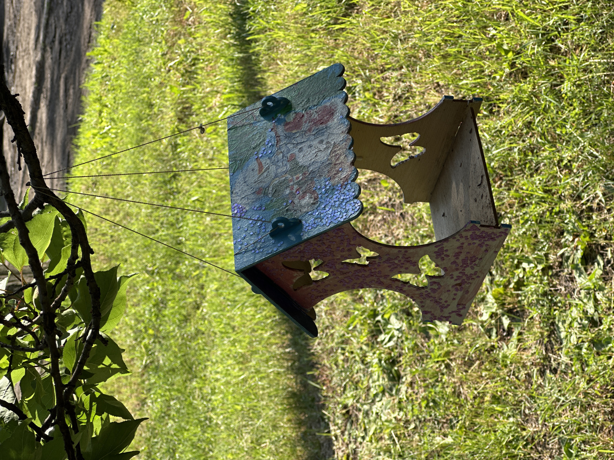 This media file is a close-up, eye-level shot of a wooden birdhouse hanging from thin wires against a background of green grass and a blurred, textured grey surface. The birdhouse is elaborately decorated with painted designs, including what appears to be a floral pattern with pink and blue hues, and scattered small, shiny blue specks. The sides of the birdhouse have decorative cut-out shapes resembling butterflies. Two dark blue, possibly glass or plastic, decorative elements are attached to the painted panels. The birdhouse is suspended from thin, dark wires that are visible running upwards and to the left. In the foreground on the left, a leafy green branch from a tree is partially in focus, with dark, textured bark and leaves casting shadows. The lighting suggests it is daytime, with sunlight casting shadows and highlighting the textures of the grass and the birdhouse. The overall impression is a charming, handcrafted decorative object in an outdoor setting. No people or text are visible in the image.
