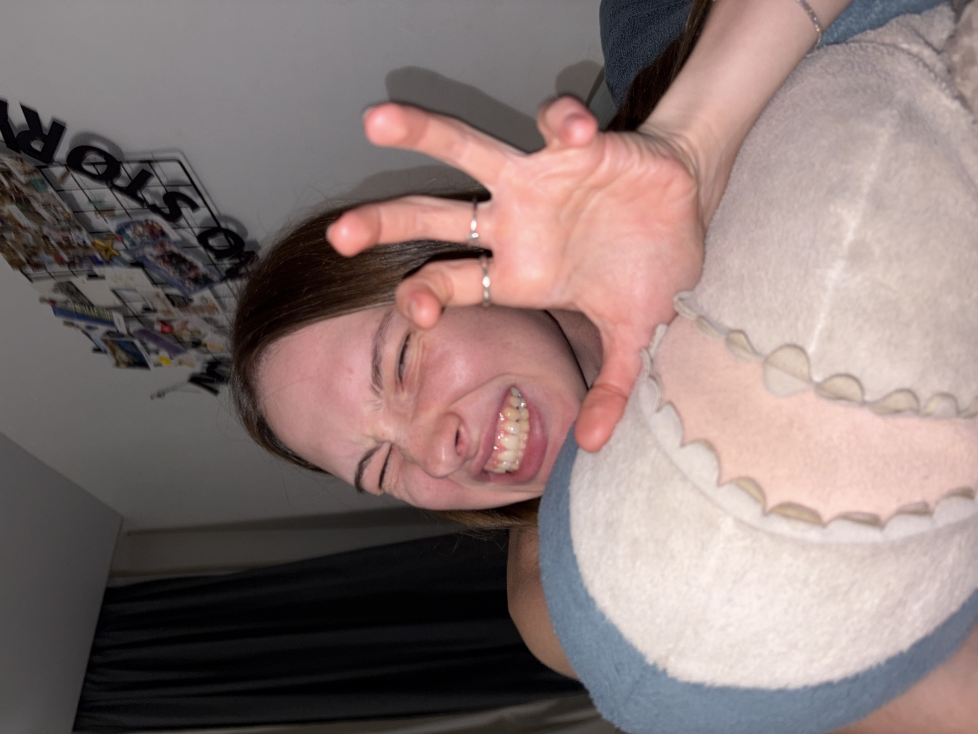 The image shows a young Caucasian female with her eyes squeezed shut and teeth bared, appearing to make a playful or menacing face. She is holding a plush toy, possibly a shark, in front of her chest with her right hand. Her left hand is raised, fingers splayed, wearing two rings. The background consists of a dark curtain on the left, a light-colored wall with a grid displaying photos and the words "NO STORY" on the right. There is no visible indication of the specific location within the city of Perm, Russia. The lighting suggests an indoor setting, likely a bedroom.