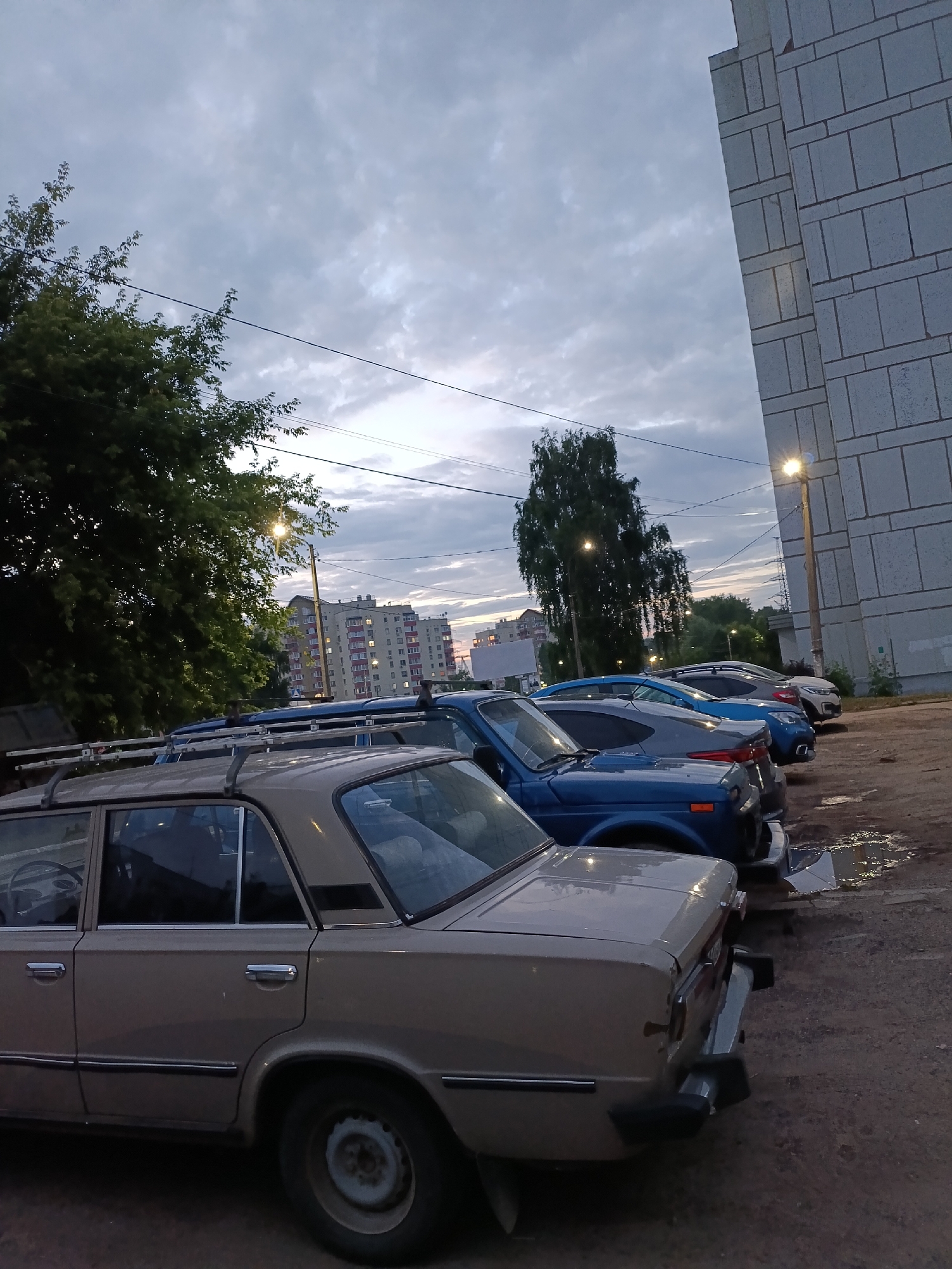 The image shows a row of parked cars in an apartment complex parking lot in Chaikovsky, Russia. A light beige sedan, possibly a Lada, is in the foreground, with its rear portion visible. Behind it are several other cars, including a blue sedan, a gray sedan, and a white SUV. The cars are parked on a dirt and gravel surface with some puddles, indicating recent rain. To the right of the parking lot is a large, light-colored apartment building with visible windows. The sky above is cloudy and overcast, suggesting it is either early morning or late evening. Tall trees are visible on the left side of the image, and apartment buildings can be seen in the background. A street lamp is illuminated near the apartment building. No people are visible in the image.